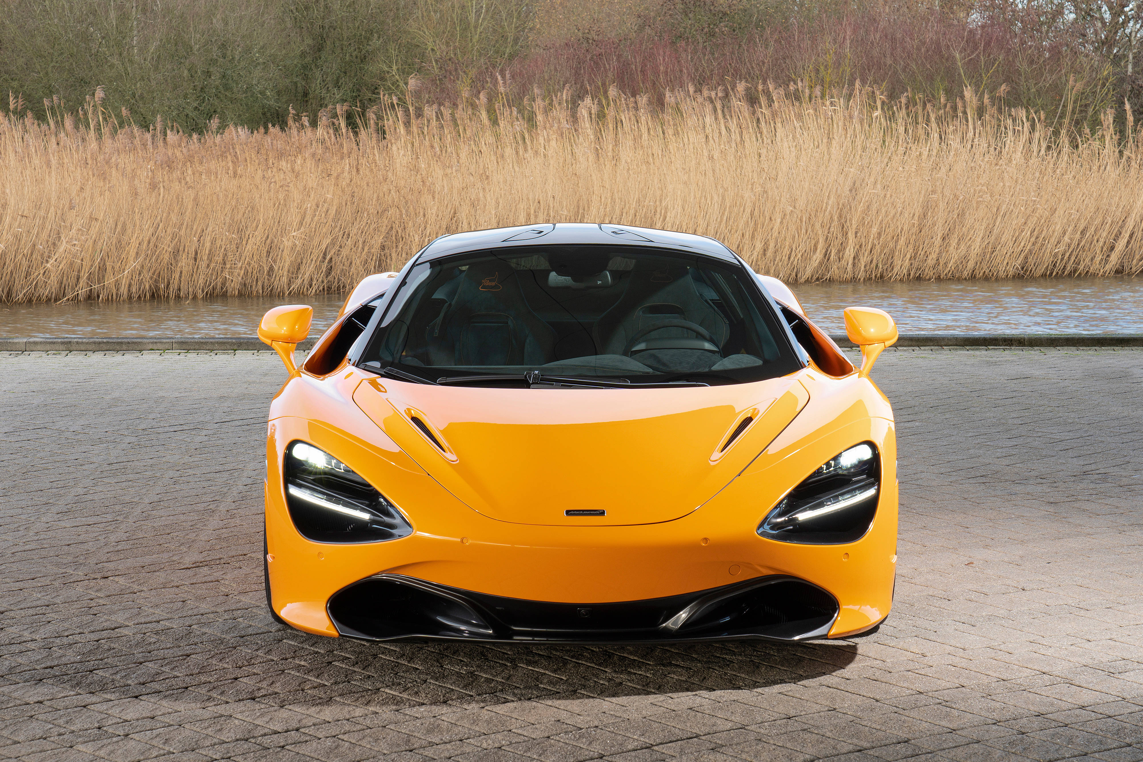 亮黃色麥拉倫720S 壁紙 — 一輛充滿活力的橙色McLaren跑車面向前方，停在鋪砌的小徑上，旁邊是平靜的水道，兩旁長滿高高的蘆葦。