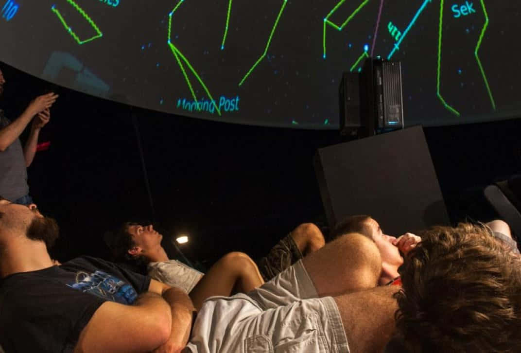 Planetarium 桌布 壁紙 — 人們仰臥著觀看一個大型弧形屏幕，屏幕上顯示發光的綠色星座和星星名稱，房間昏暗。