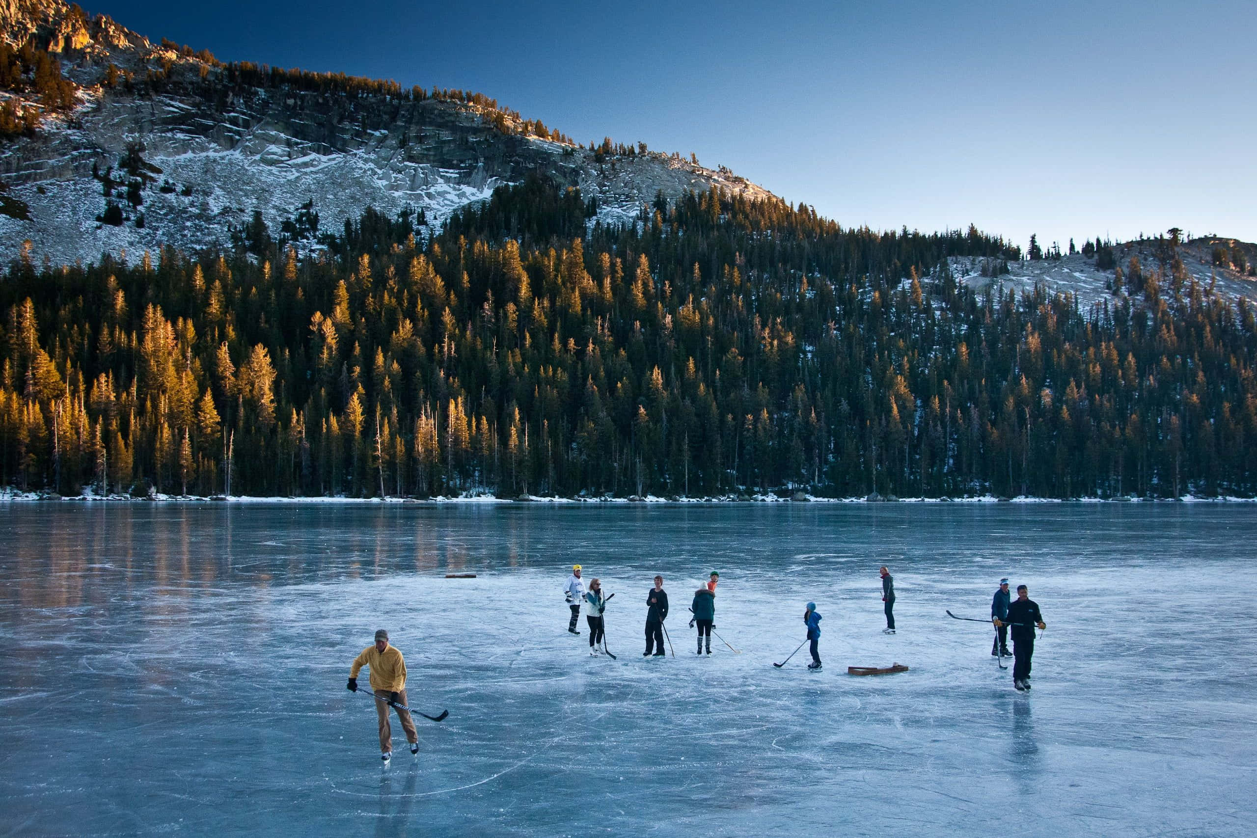 Frozen Lake 桌布 壁紙 — 人們在日落時分，在被雪覆蓋的山脈和金色松林環繞的冰凍湖面上打冰球。