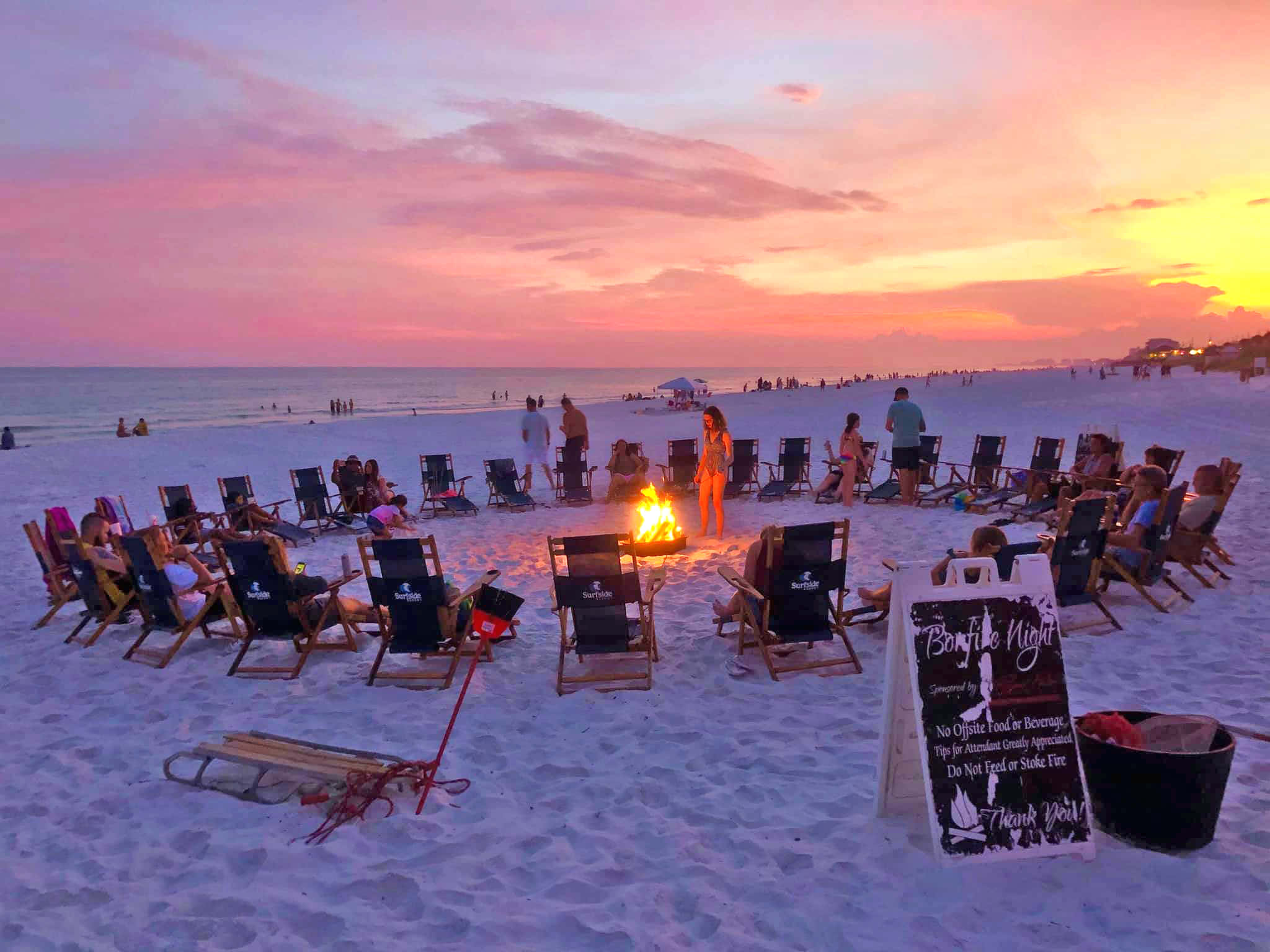 Beach Bonfire 桌布 壁紙 — 人們在日落時分聚集在海灘篝火旁，天空呈現粉橙色，海洋地平線清晰可見。