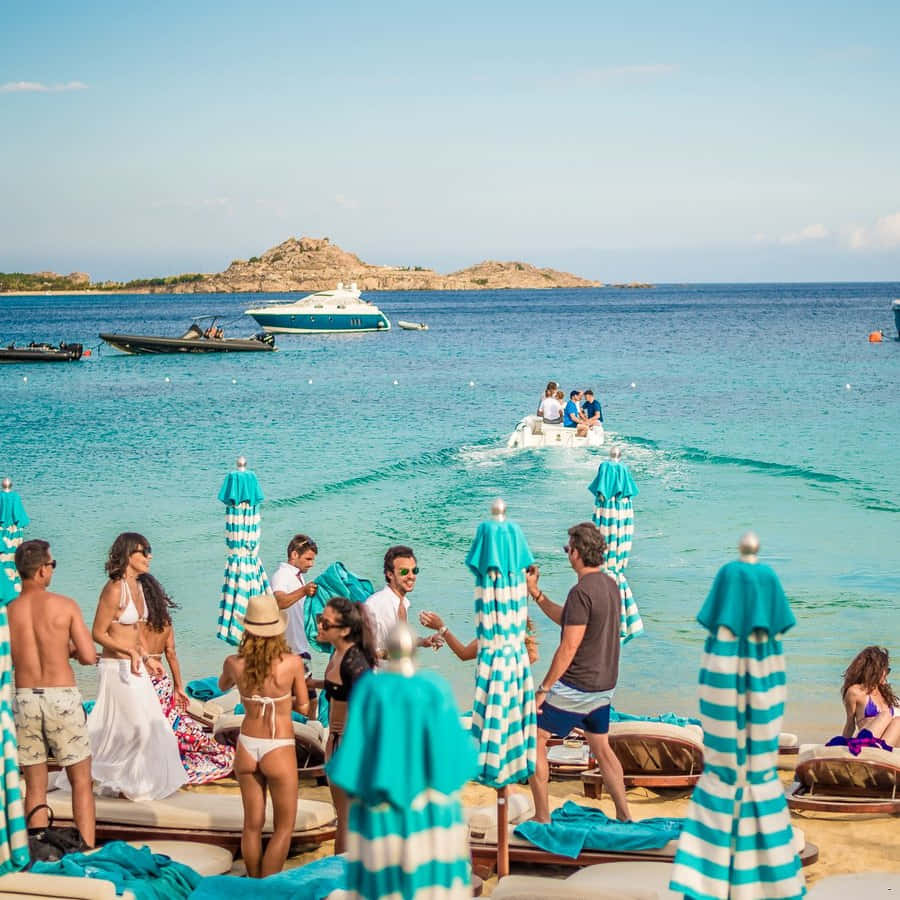 Mediterranean Beach 桌布 壁紙 — 人們在陽光明媚的海灘上，在條紋遮陽傘下放鬆，船隻在岩石島嶼附近的碧綠海面上滑行。