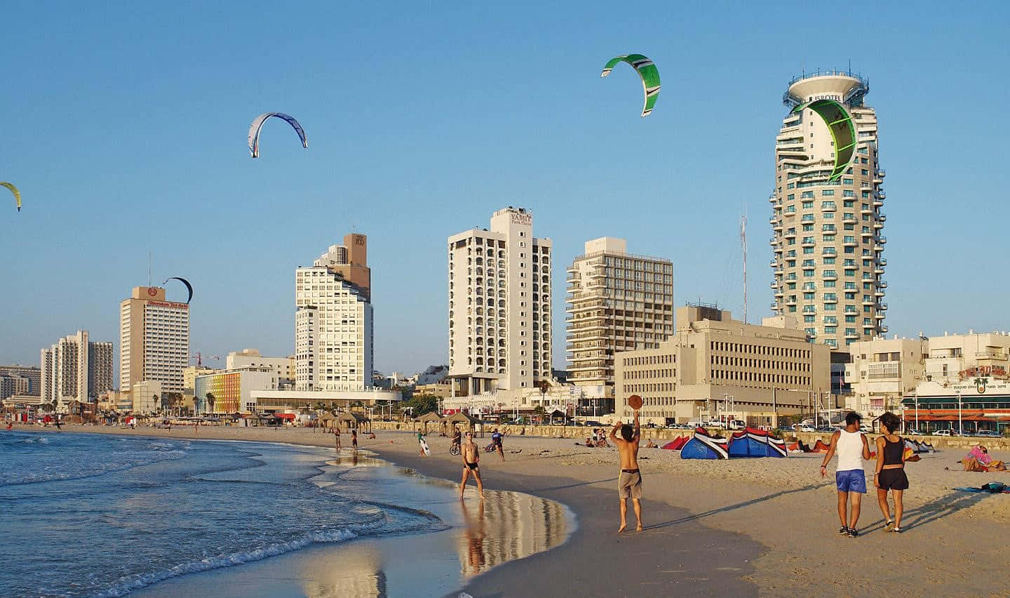 Israel 桌布 壁紙 — 人們在陽光明媚的海灘上享受，風箏衝浪者放飛色彩繽紛的風箏，背景是現代城市摩天大樓。