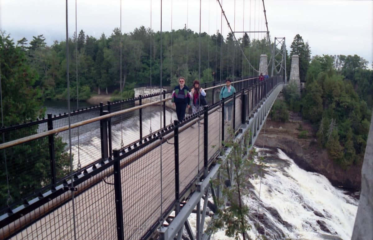Fall Bridge 桌布 壁紙 — 人們走過一座金屬吊橋，橋下是湍急的河流，周圍是茂密的綠色森林，天空多雲。