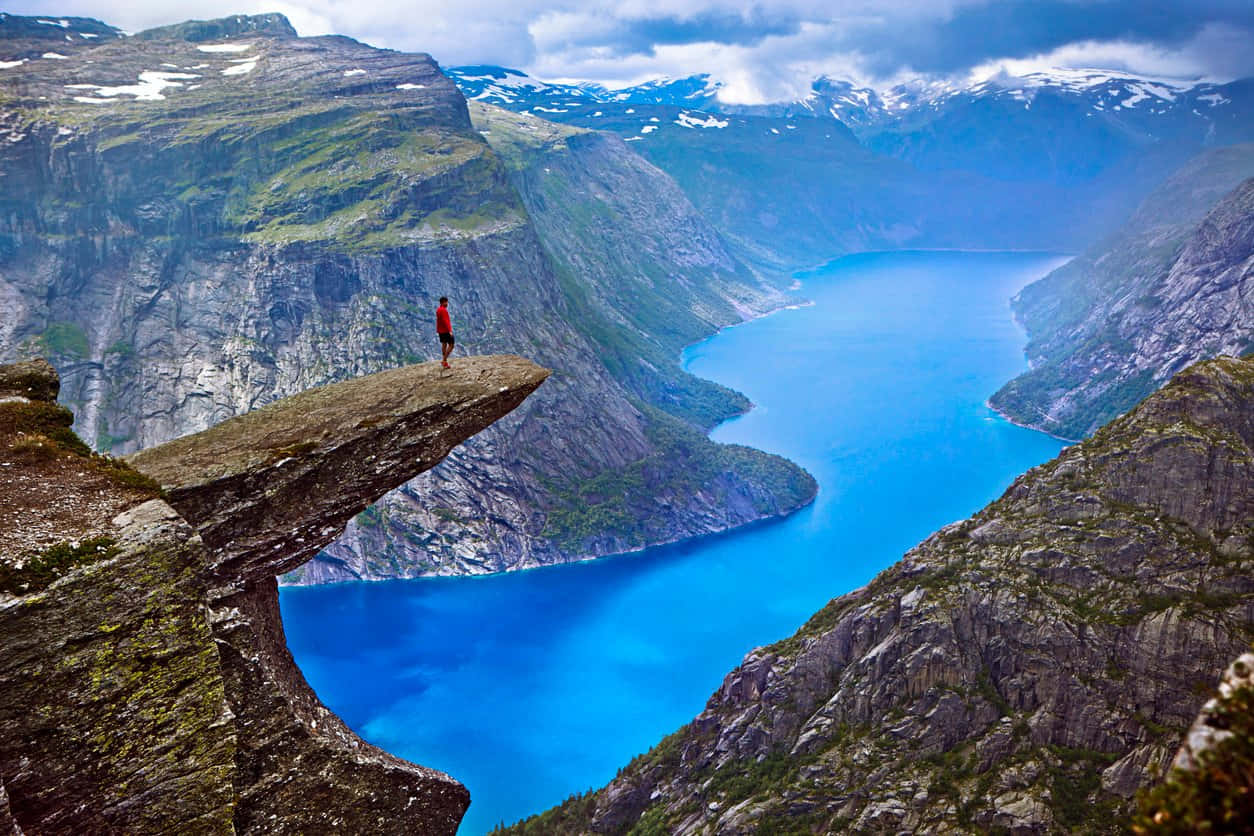 人在Trolltunga岩石上俯瞰峽灣 壁紙 — 一個孤獨的紅色人影站在巨大的懸崖頂上，俯瞰著夾在雄偉的綠色和積雪山脈之間、在陰天天空下的 vibrant 藍色峽灣。