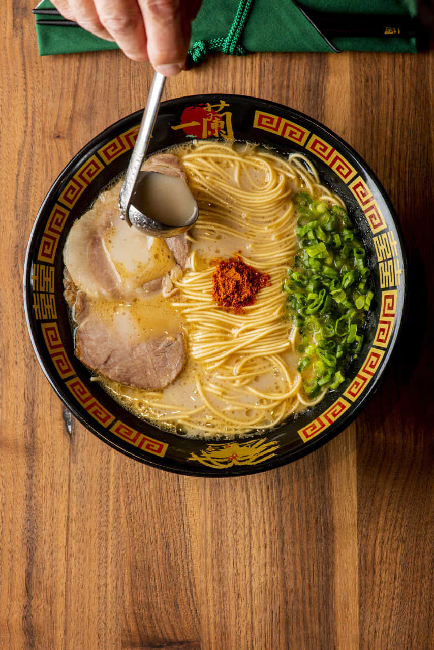 人物手抓拉麵 壁紙 — 一碗冒煙的拉麵，有麵條、湯底、豬肉片、蔥花和辣椒油，正被湯匙舀起。