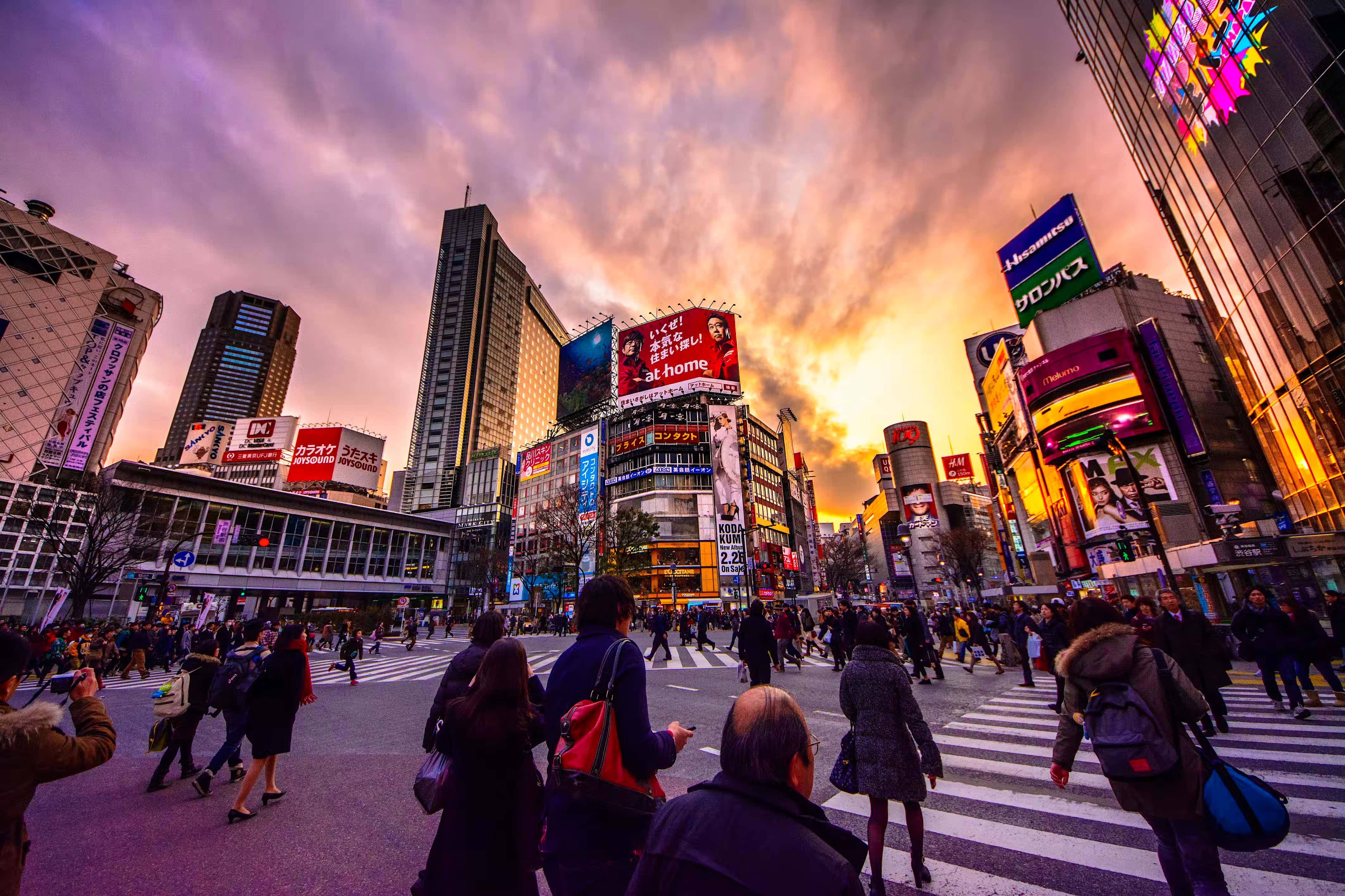 東京 桌布 壁紙 — 人群在日落時分穿過繁華的東京街道，霓虹燈在火紅的紫色和橙色天空下閃耀。