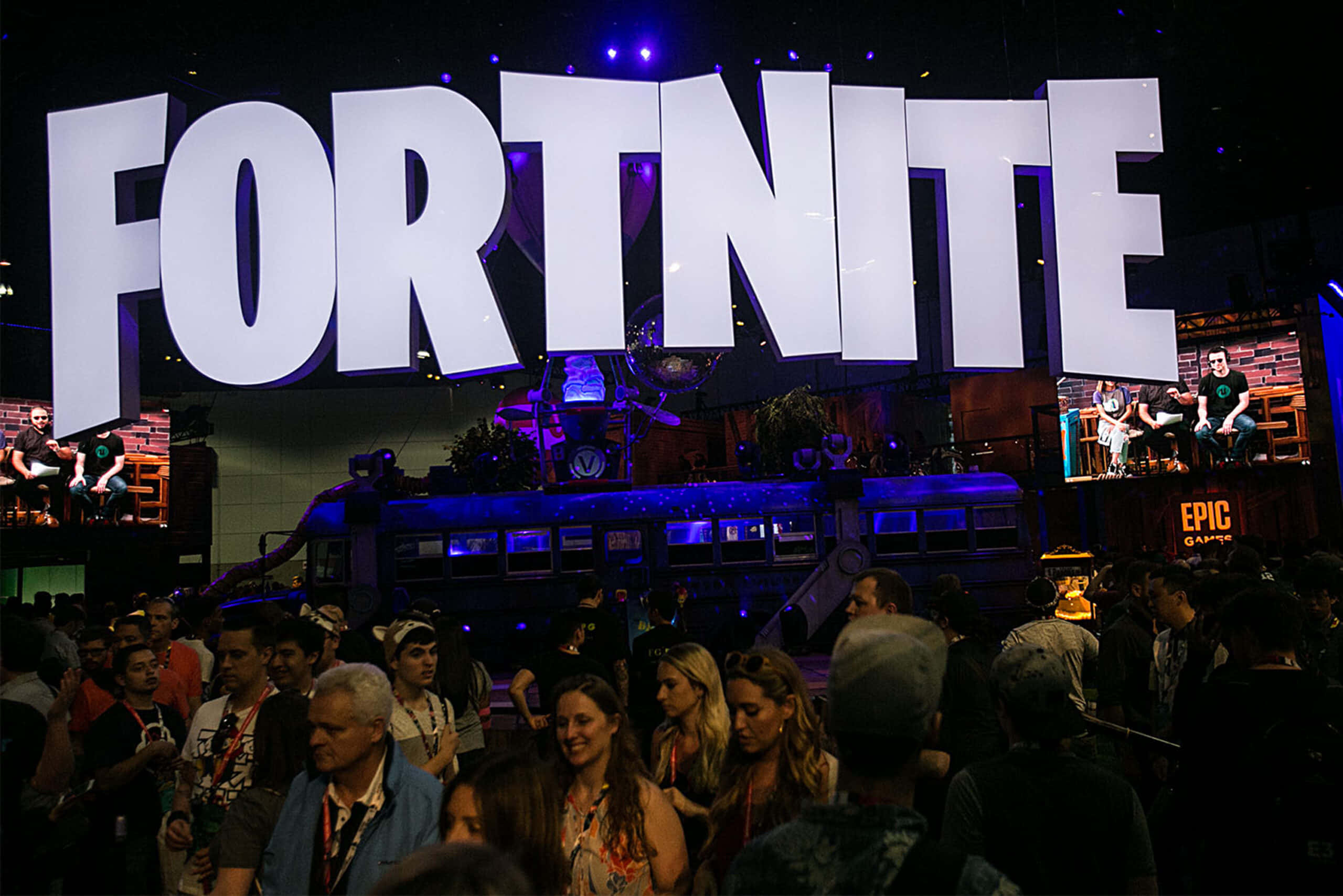 Gaming Conventions 桌布 壁紙 — 人群在昏暗的環境燈光下，聚集在遊戲大會上一個巨大的發光 Fortnite 標誌下方。