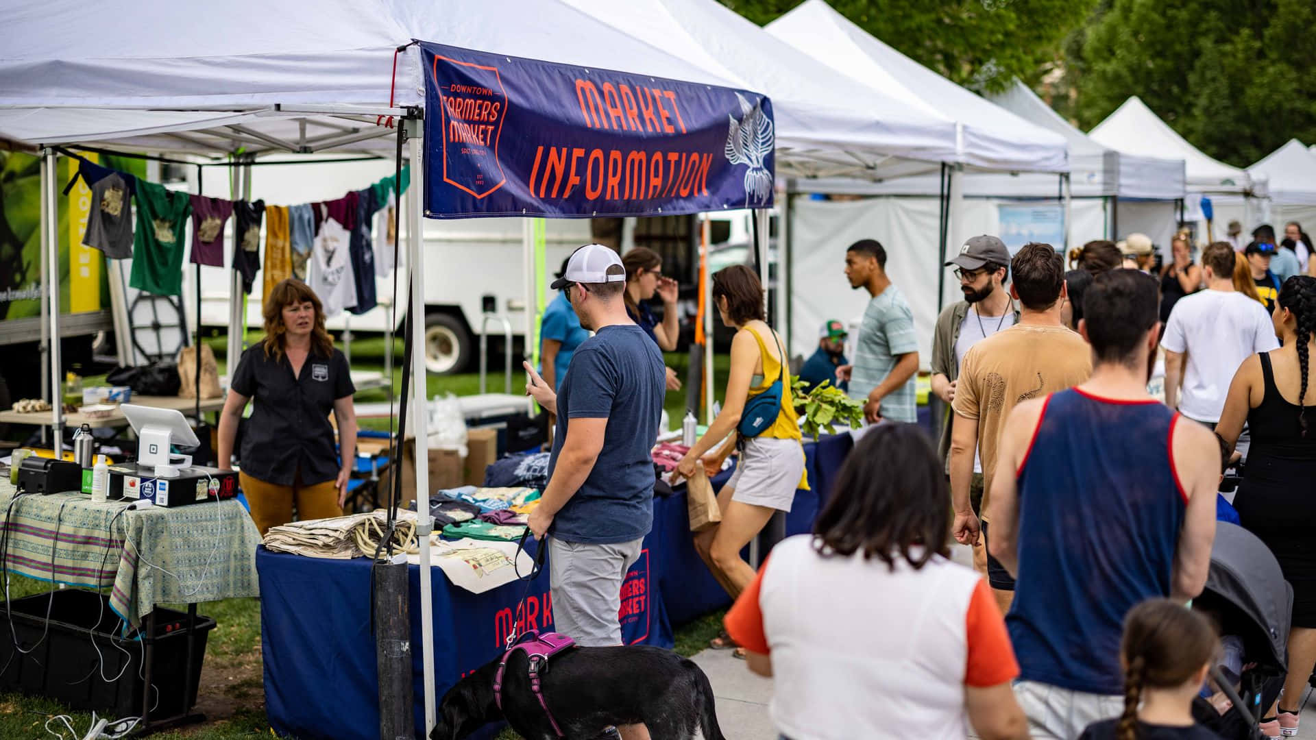 Farmers Market 桌布 壁紙 — 人群在晴朗的日子裡，在白色遮陽篷下瀏覽熱鬧的戶外農貿市場的攤位。