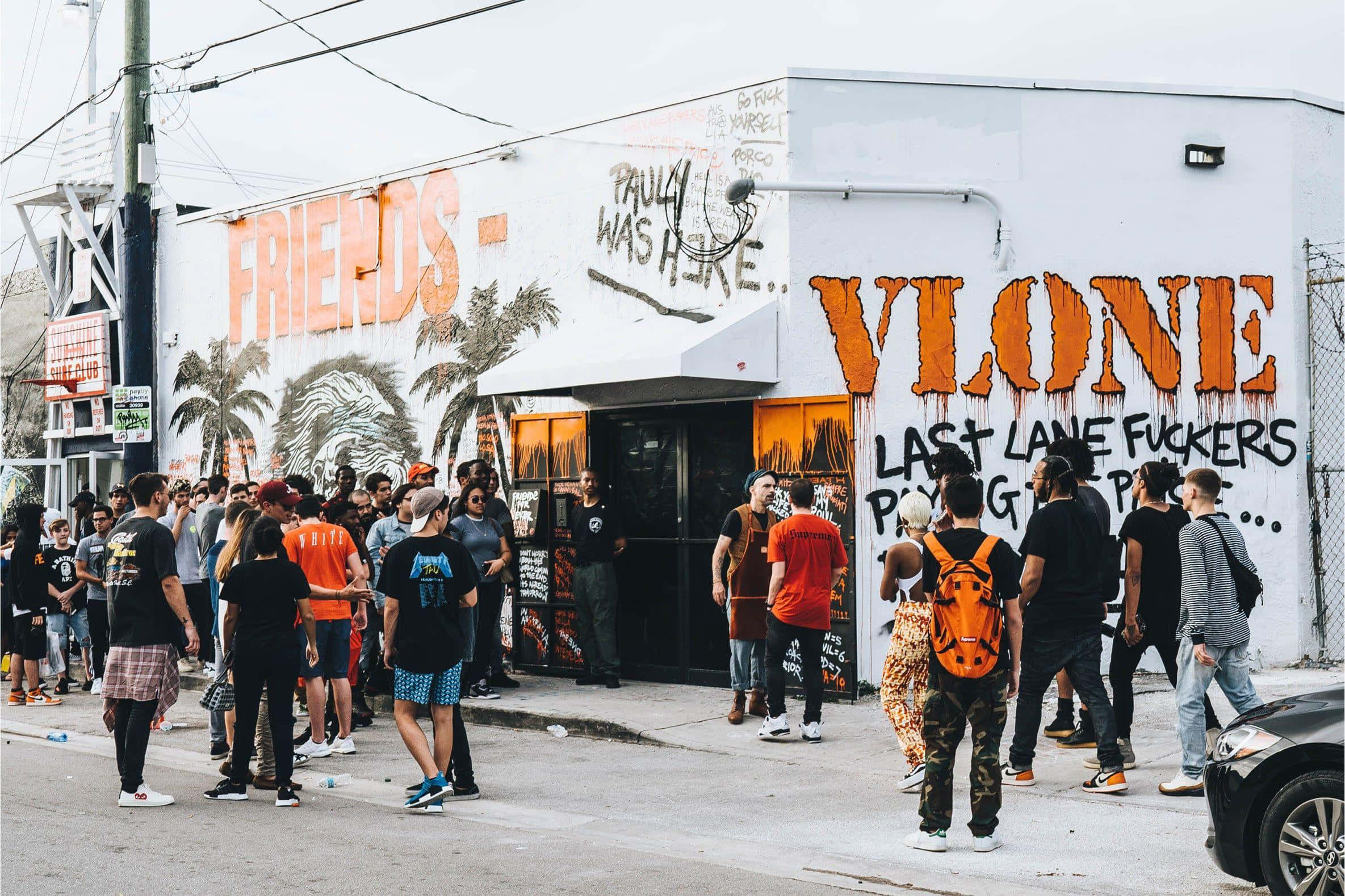 Vlone 桌布 壁紙 — 人群聚集在一座被塗鴉覆蓋的建築物外，建築物上有大膽的橙色和黑色街頭藝術，天空多雲。