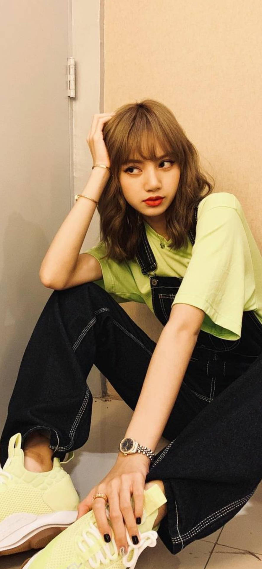 仁川機場時尚Lisa BLACKPINK 高畫質照片 壁紙 — 一位年輕女性留著波浪長髮，身穿青檸綠色襯衫和黑色吊帶褲，搭配同款運動鞋，隨意地靠牆而坐。