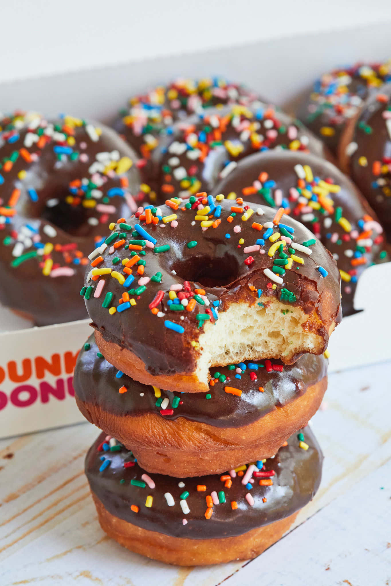 今天就享用Dunkin' Donuts的甜蜜點心吧！ 壁紙 — 一堆朱古力糖霜冬甩，上面有彩色糖針，其中一個被咬了一口，露出軟綿綿的內部。