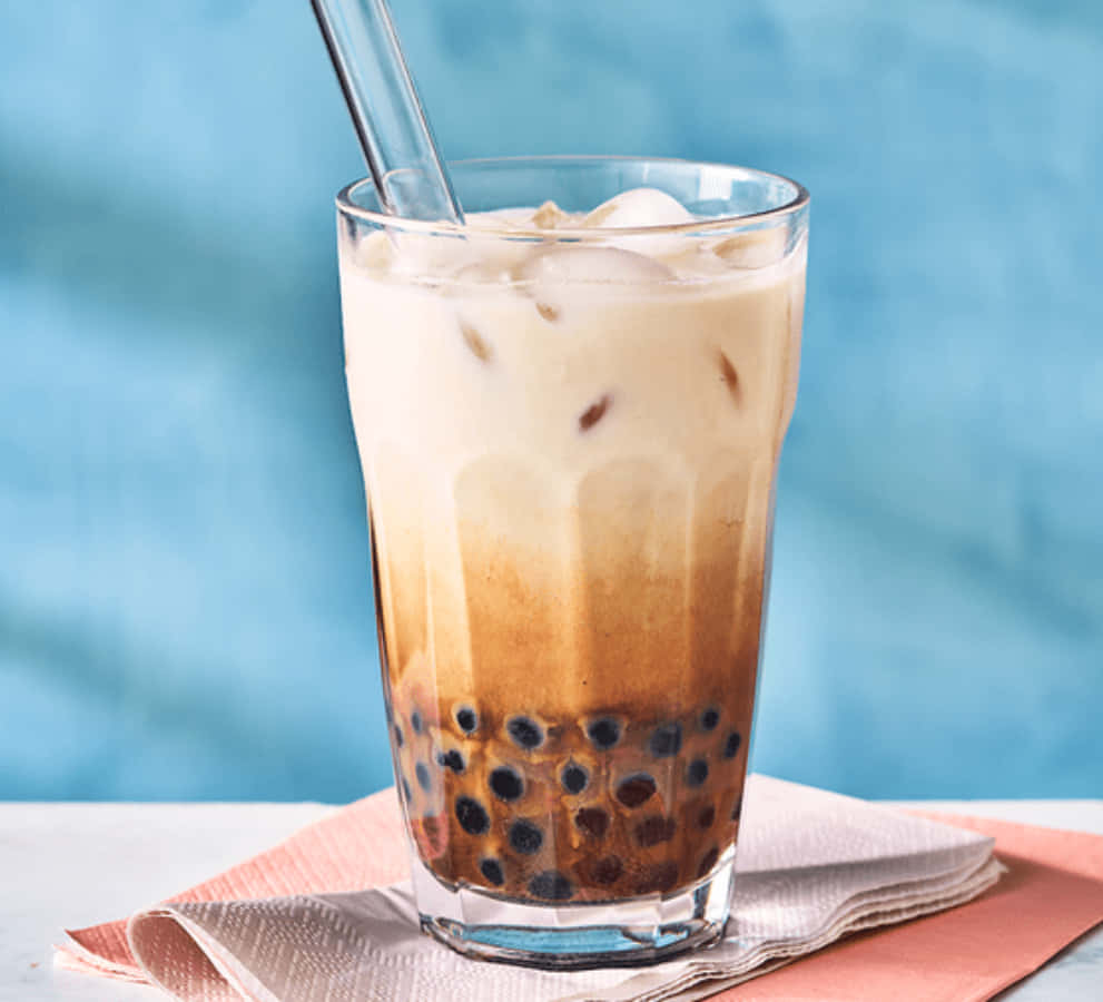 今天就享用一杯美味的波霸奶茶吧 壁紙 — 一杯高身凍咖啡，內有黑色珍珠和冰塊，放在摺疊的粉紅色餐巾上，背景是柔和的藍色。