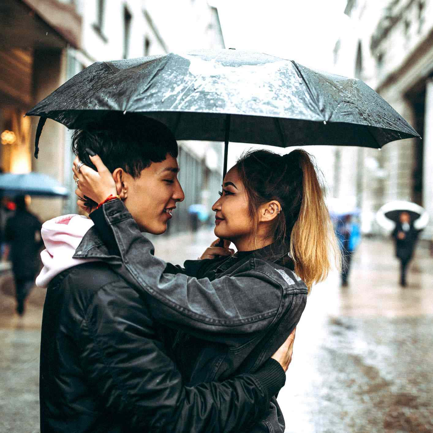 他們在雨中共享了一個親密時刻 壁紙 — 一對微笑的情侶在雨天的城市街道上，在黑色雨傘下擁抱，濕漉漉的人行道和模糊的行人。
