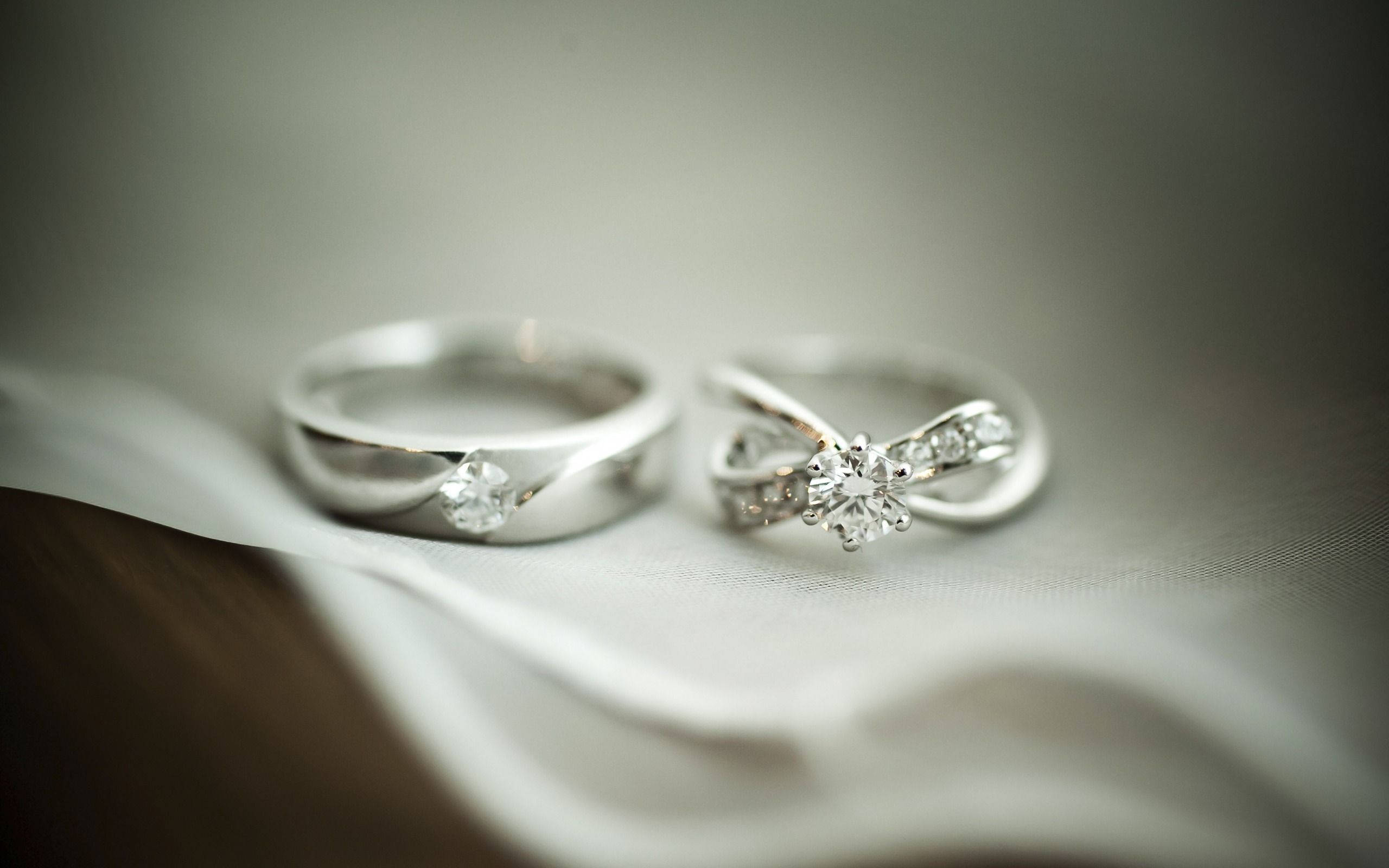 他的與她的銀婚戒 壁紙 — 兩枚優雅的鑽石結婚戒指放在柔軟的白色布料上，景深較淺。
