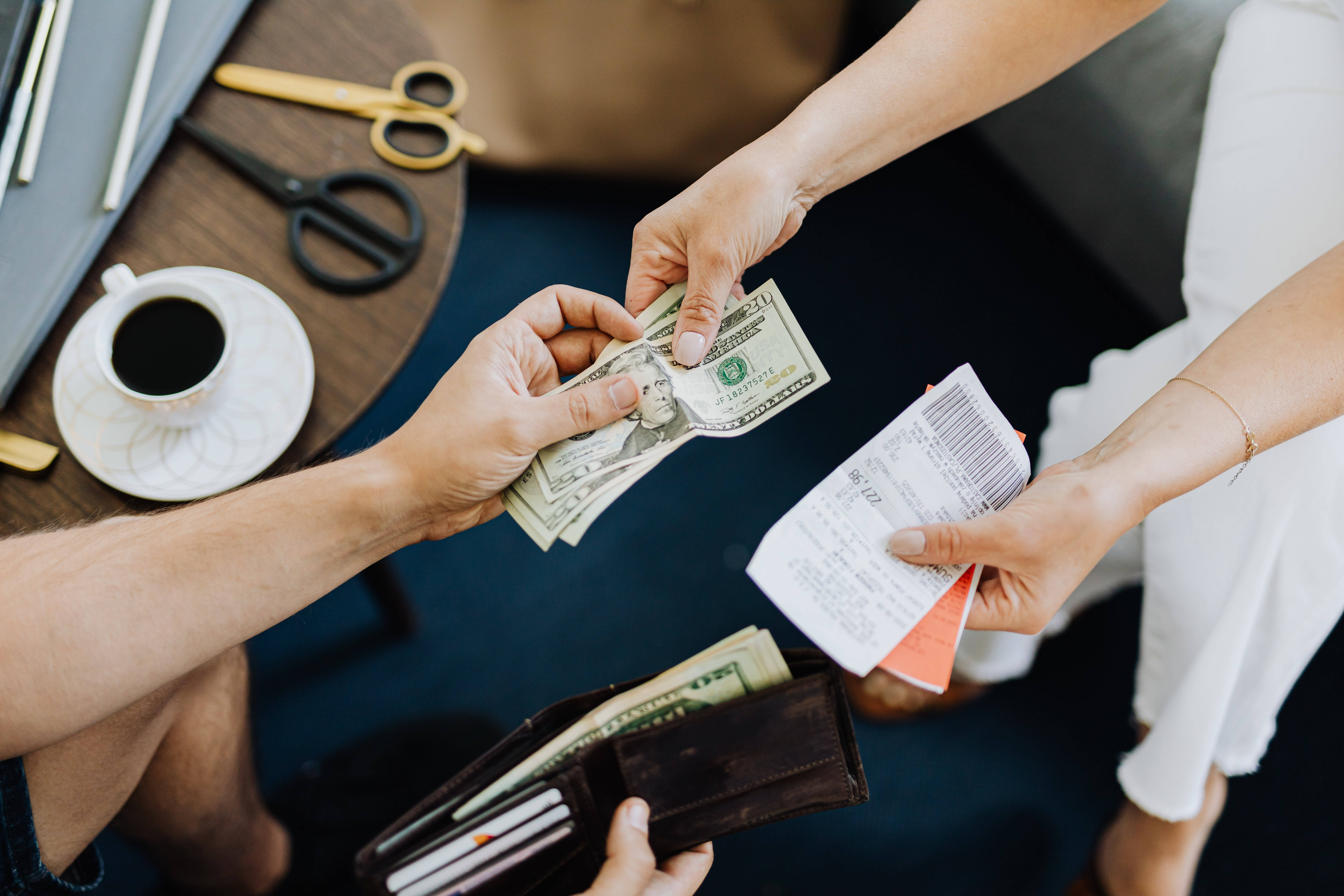 付款與收據 壁紙 — Overhead view of hands exchanging cash and receipts at a wooden table with coffee and scissors nearby.
