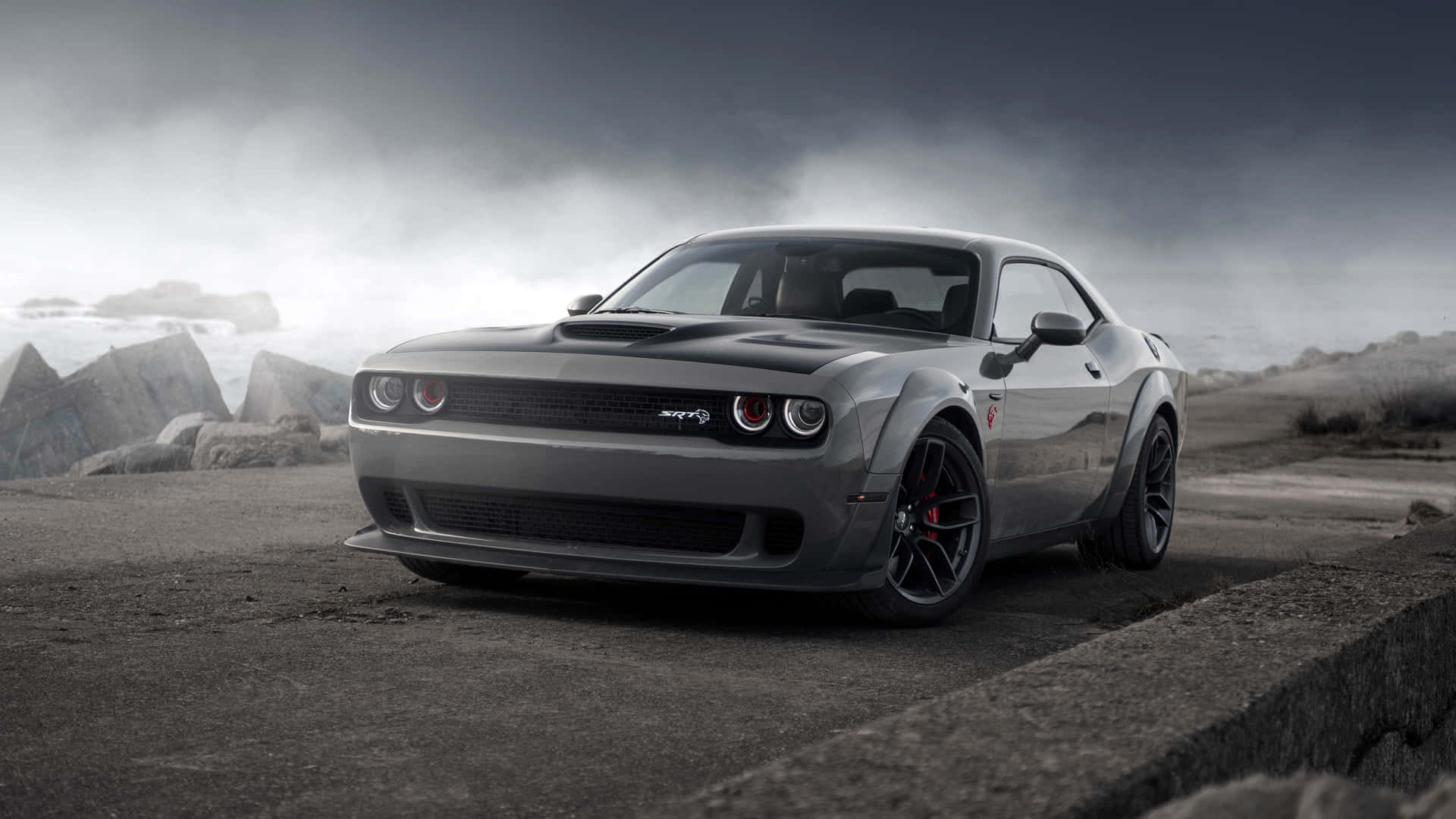 灰色 Dodge Challenger SRT Hellcat 在混凝土邊緣上，背景有風暴天空和岩石。