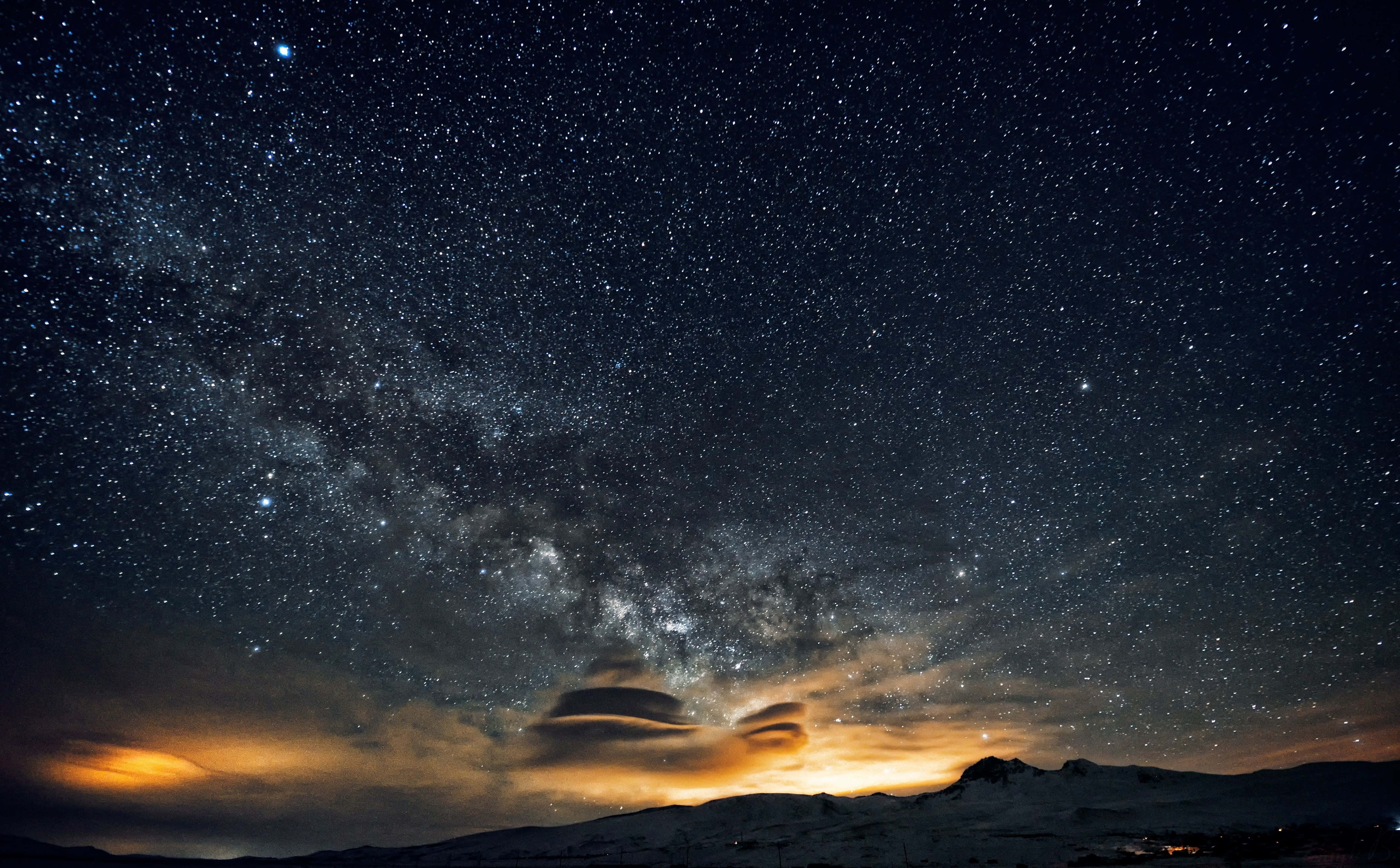 Awe-inspiring 桌布 壁紙 — 令人嘆為觀止的夜空，佈滿無數繁星，銀河在積雪的山峰上拱起，下方是發光的橙色雲層。