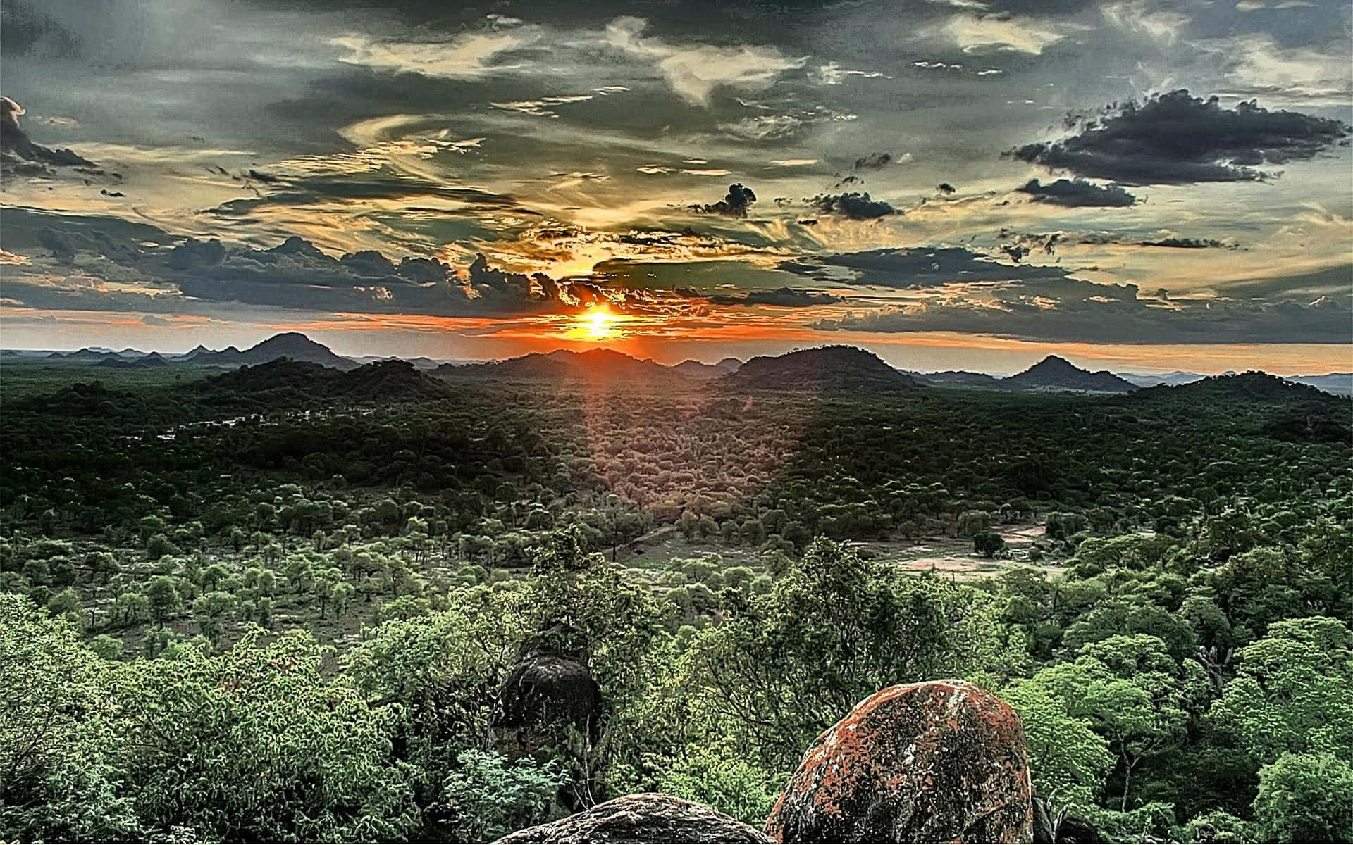 African Landscapes 桌布 壁紙 — 令人嘆為觀止的日落將天空染成火熱的橙色和金色，落在遠山之後，在下方茂密的綠色森林上投下長長的陰影。
