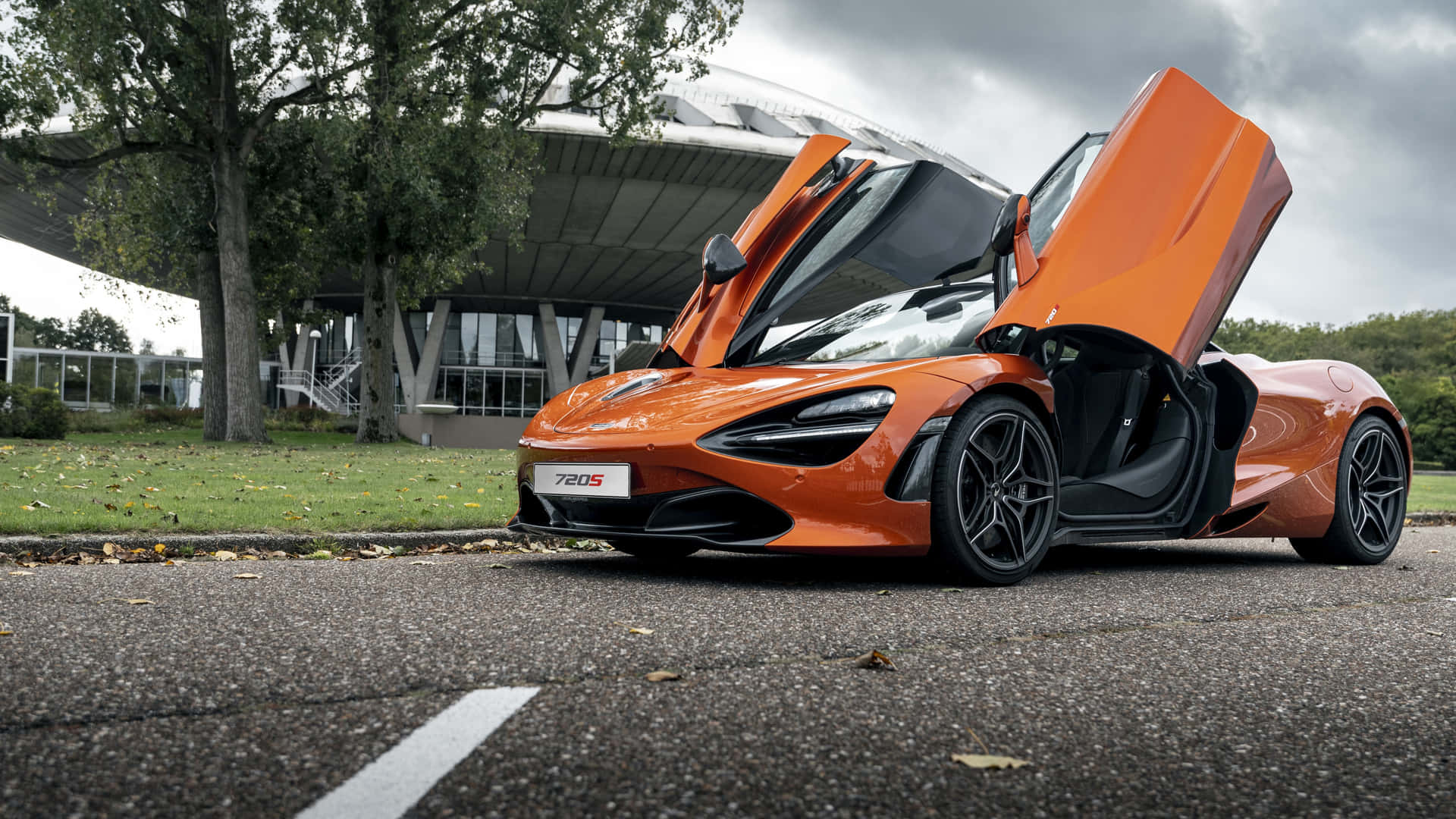 橙色McLaren 780S超級跑車車門打開，停泊在戶外，附近有樹木和建築物。