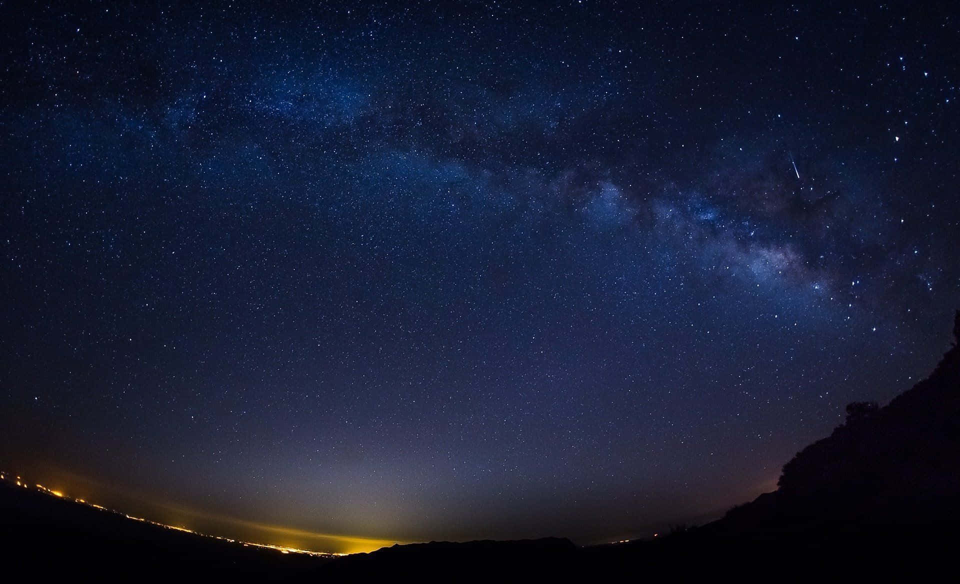 星空 桌布 壁紙 — 令人屏息的夜空，充滿無數星星和發光的銀河，在黑暗的山丘輪廓上拱起，下方是遠處的城市燈光。
