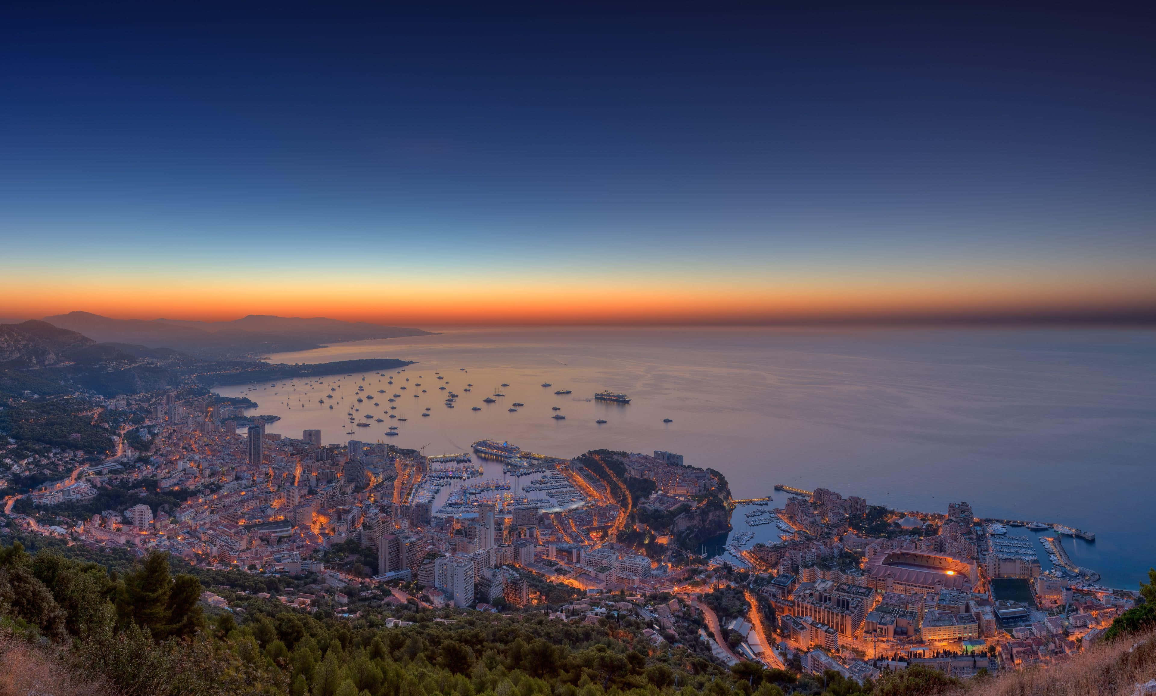 令人屏息的摩納哥海濱景觀 壁紙 — 摩納哥日落時的全景，閃爍的城市燈光倒映在平靜的海面上，近岸停泊著船隻。