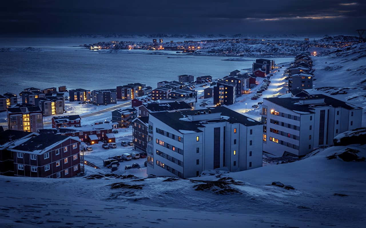 令人屏息的格陵蘭風景 壁紙 — 積雪覆蓋的建築物在俯瞰冰封海洋的黑暗冬日天空中溫暖地發光。