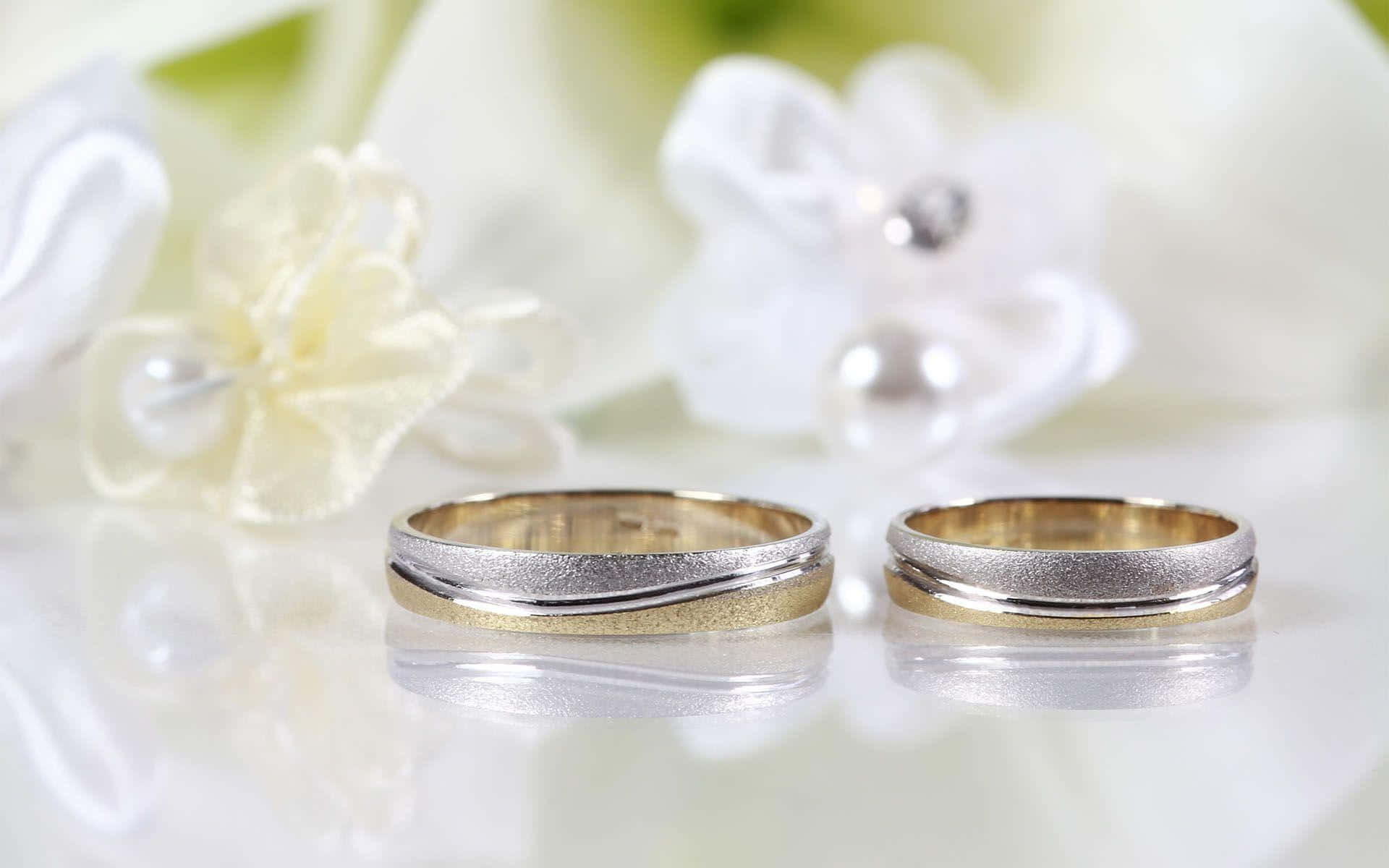 令人屏息的銀色婚戒圖像 壁紙 — 兩枚優雅的金色和銀色結婚戒指放在反光表面上，旁邊有柔軟的白色花卉裝飾。