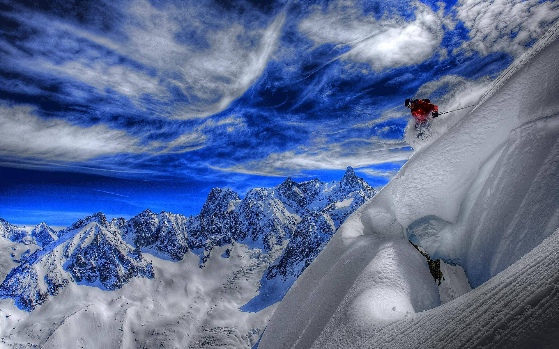 令人屏息的雪山滑雪風景 壁紙 — 一名身穿紅色滑雪服的滑雪者在陡峭的山脊上穿過深粉雪，背景是戲劇性的藍天和縷縷白雲。
