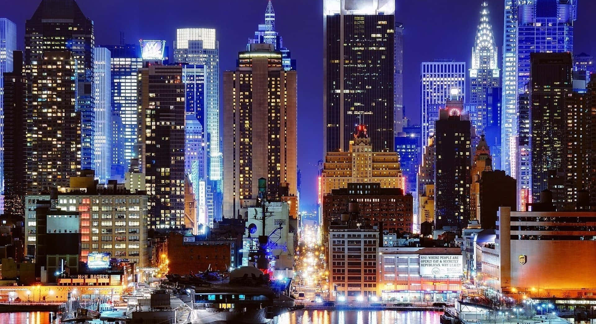 紐約 桌布 壁紙 — 令人目眩的夜景城市景觀，高聳的摩天大樓閃爍著藍色和金色的燈光，倒映在水面上。