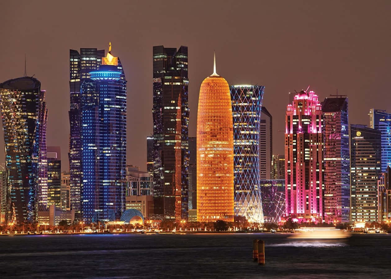 Qatar 桌布 壁紙 — 令人眼花繚亂的夜景，現代摩天大樓閃爍著充滿活力的藍色、粉紅色和橙色燈光，倒映在平靜的水面上。