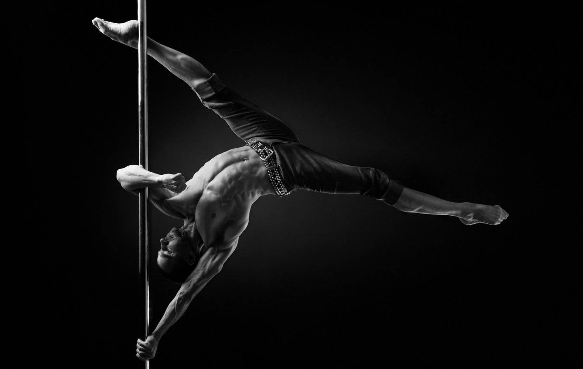 令人著迷的 Pole Dance 舞者示範 壁紙 — 一名肌肉發達的男子在純黑背景前，以高對比黑白呈現動態鋼管舞姿勢。