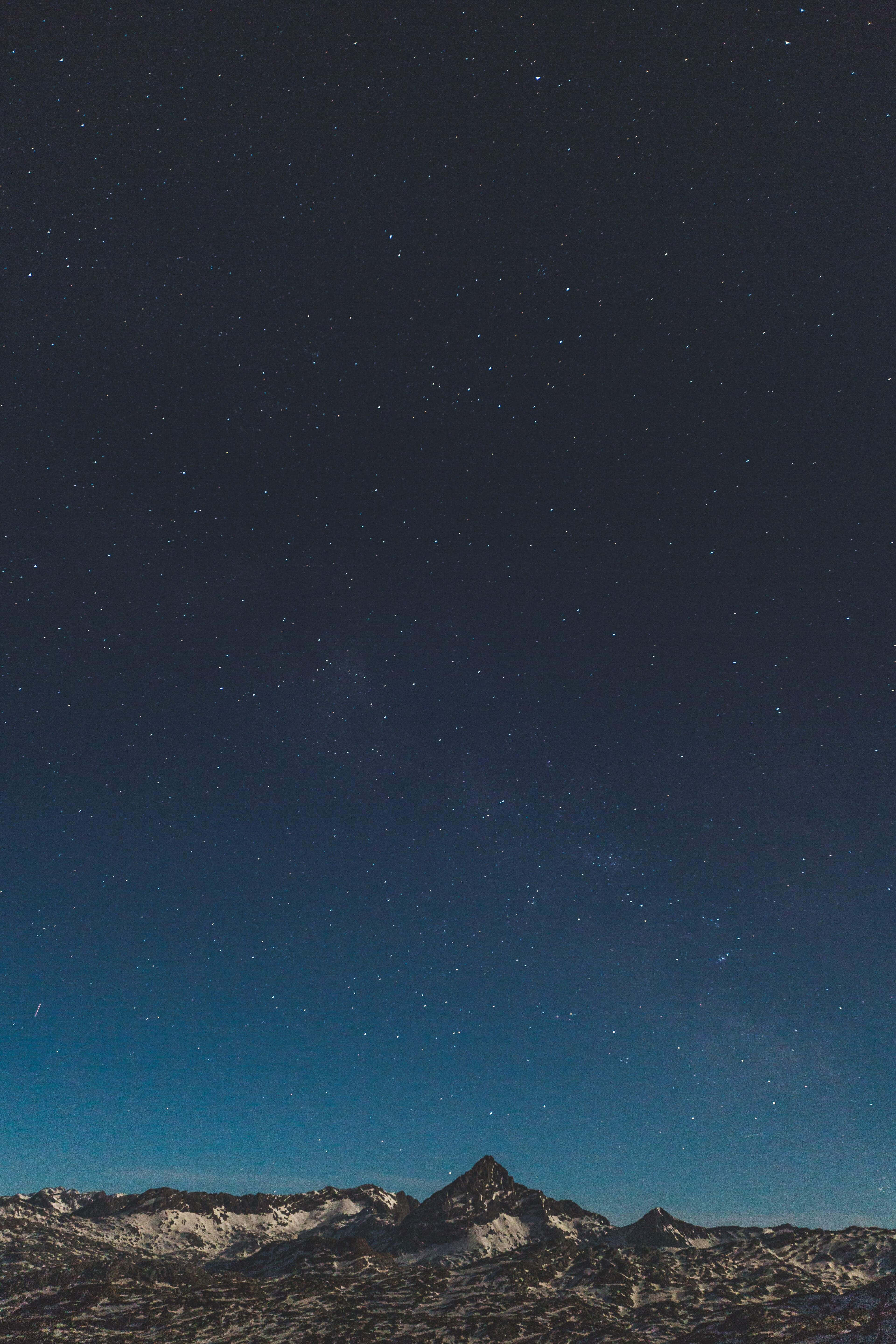 令人難以抗拒的山星 iPhone 背景 壁紙 — 白雪覆蓋的山峰在廣闊的星空下聳立，星空閃爍著宇宙藍和深靛藍色調。