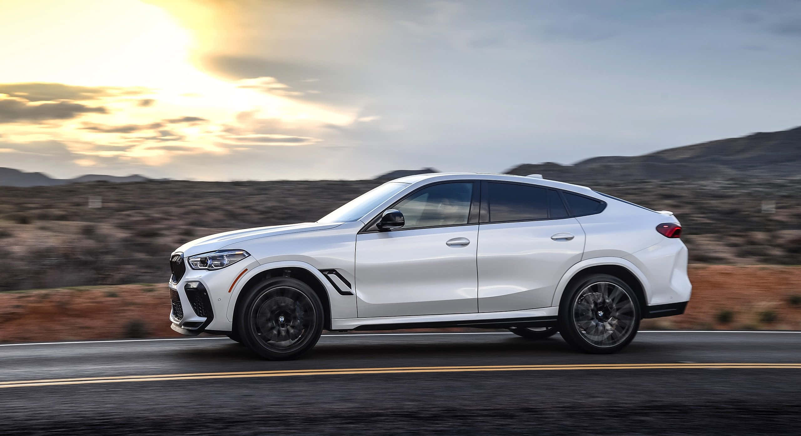令人驚嘆的BMW X6 M 壁紙 — 一輛時尚的白色BMW X6在沙漠公路上行駛，夕陽西下，金色的光芒投下長長的影子。