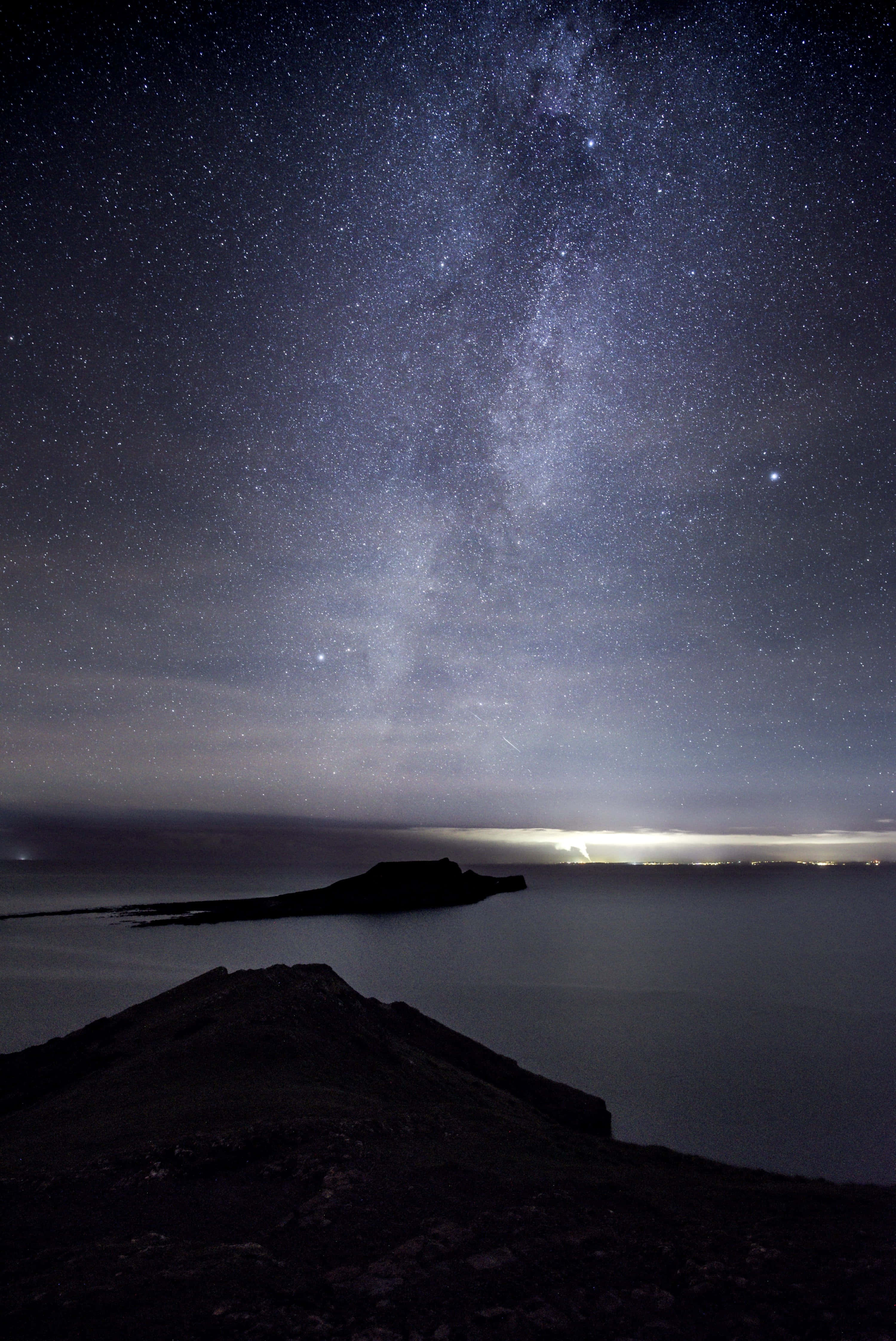 Dark Island 桌布 壁紙 — 令人驚嘆的夜空景觀，銀河在黑暗的海岸景觀上方拱起，地平線上有遠處的城市燈光。