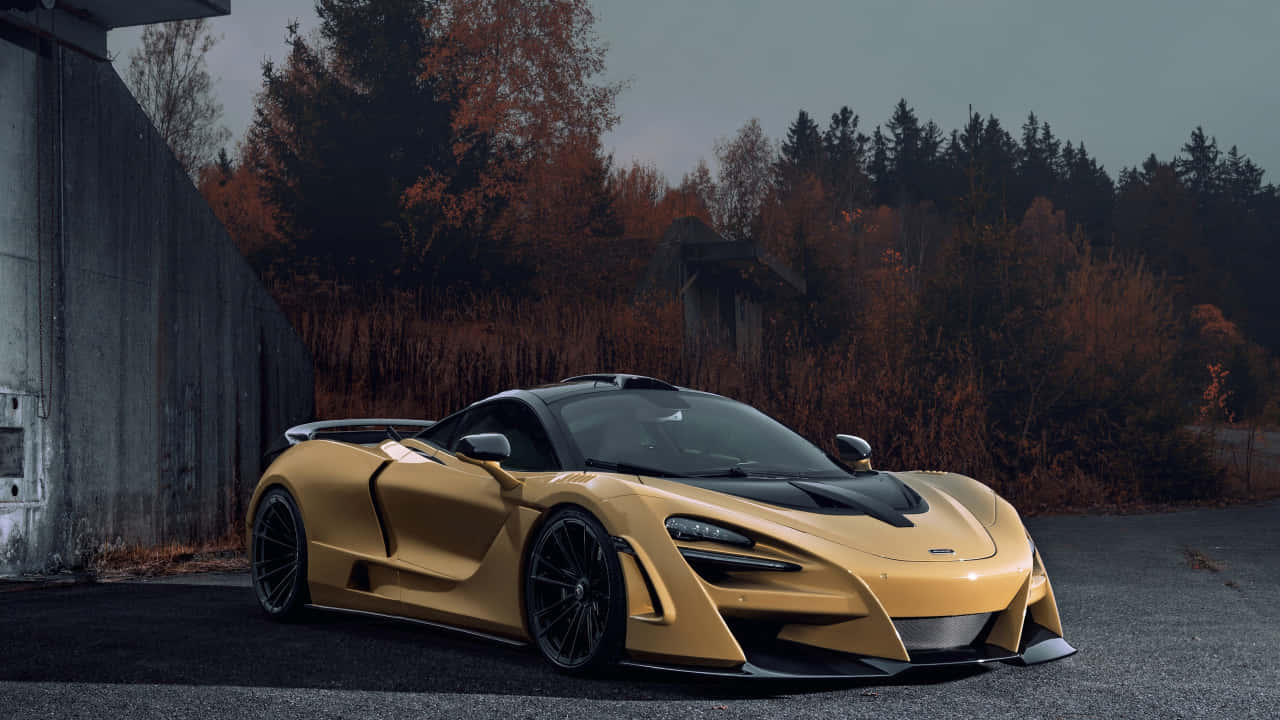 令人驚嘆的奢華與強勁麥拉倫720S 壁紙 — 一輛金色McLaren超級跑車停在瀝青路上，旁邊是混凝土牆，背景有秋天的樹木。