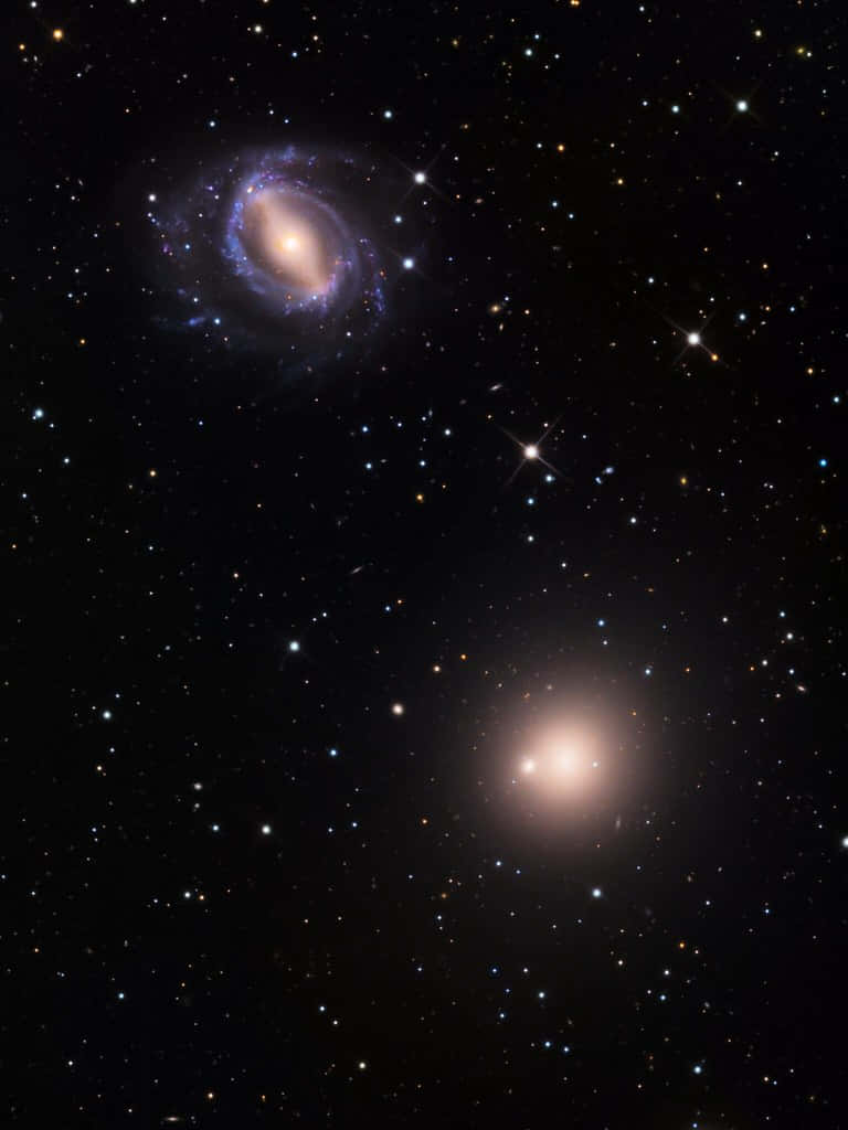Elliptical Galaxy 桌布 壁紙 — 令人驚嘆的宇宙景觀，兩個發光的星系在佈滿星星的黑色虛空中以紫色和金色色調旋轉。