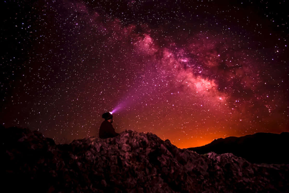 令人驚嘆的星系美景 壁紙 — 夜晚，一個孤獨的身影坐在岩石山脊上，用頭燈照向充滿活力的粉紅色和橙色銀河系。