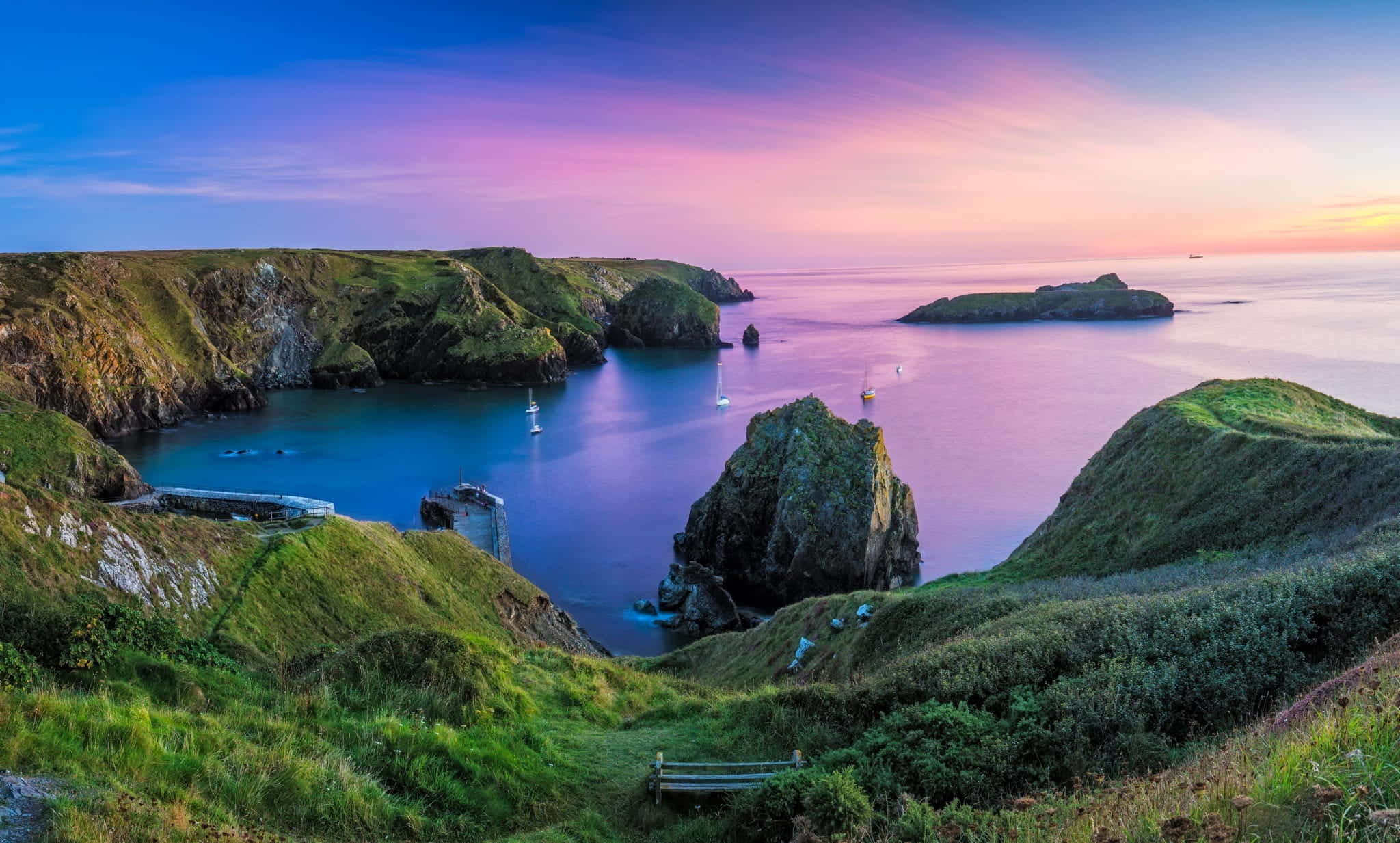 UK 桌布 壁紙 — 令人驚嘆的海岸小灣日落景色，充滿活力的紫色和粉紅色天空倒映在平靜的碧綠海水中，周圍環繞著鬱鬱蔥蔥的綠色懸崖和帆船。