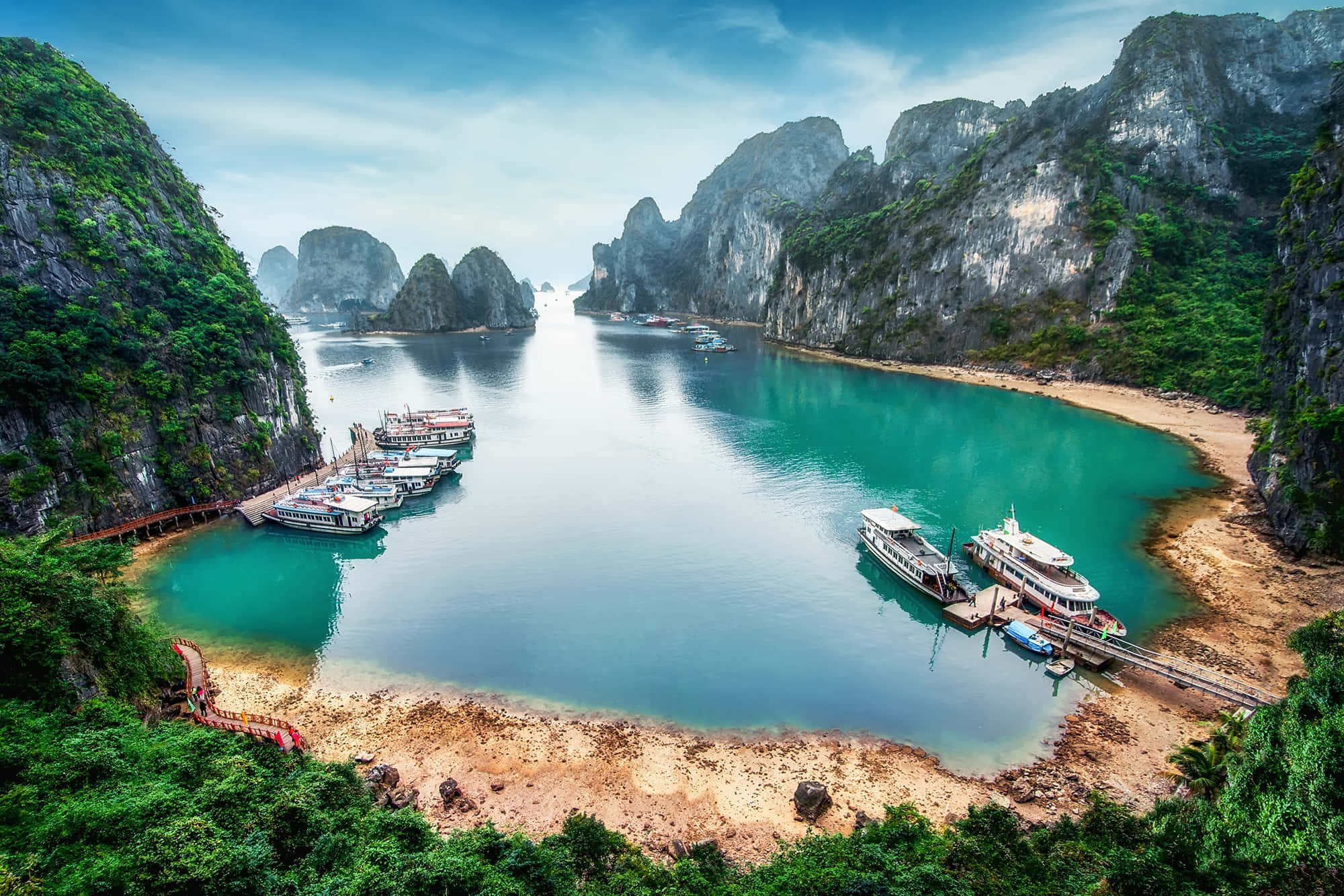 Vietnam 桌布 壁紙 — 令人驚嘆的鳥瞰圖，展現一個碧綠色的海灣，坐落在高聳的綠色石灰岩懸崖之間，沙灘上停泊著船隻。