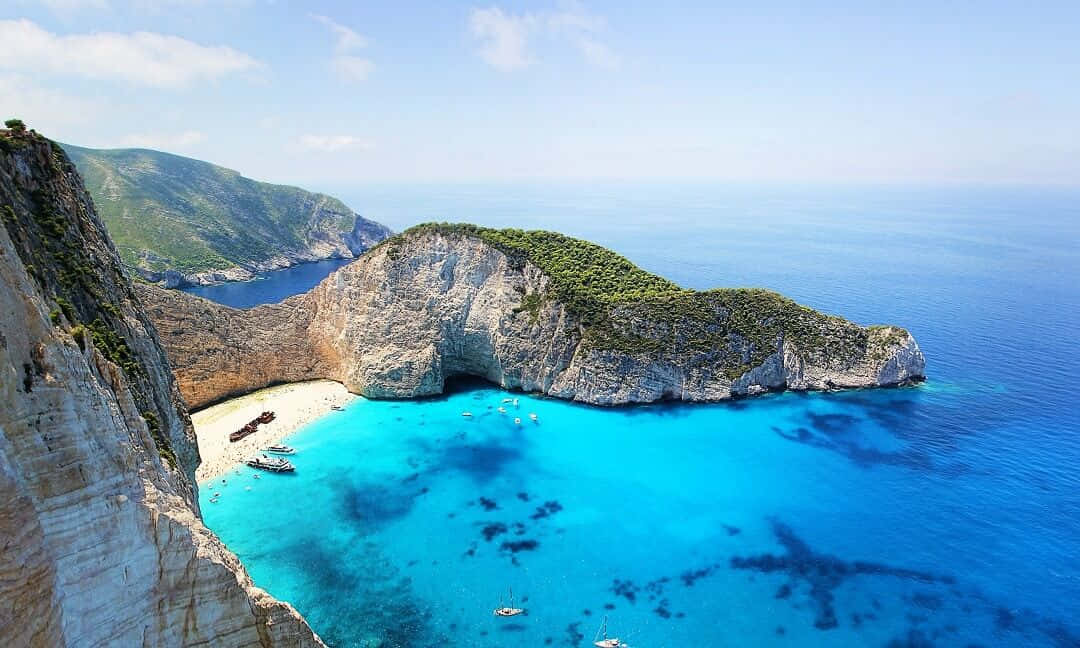 Greek Island 桌布 壁紙 — 令人驚嘆的鳥瞰圖，展現一個隱藏在高聳白色懸崖之間的僻靜碧綠海灣，船隻停泊在清澈見底的水中。