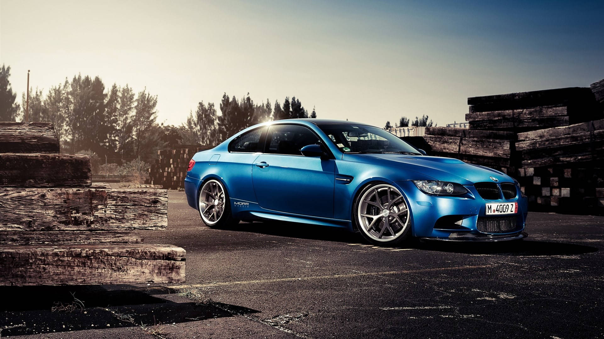 藍色BMW M3跑車停泊在堆疊木頭旁邊，夕陽西下。
