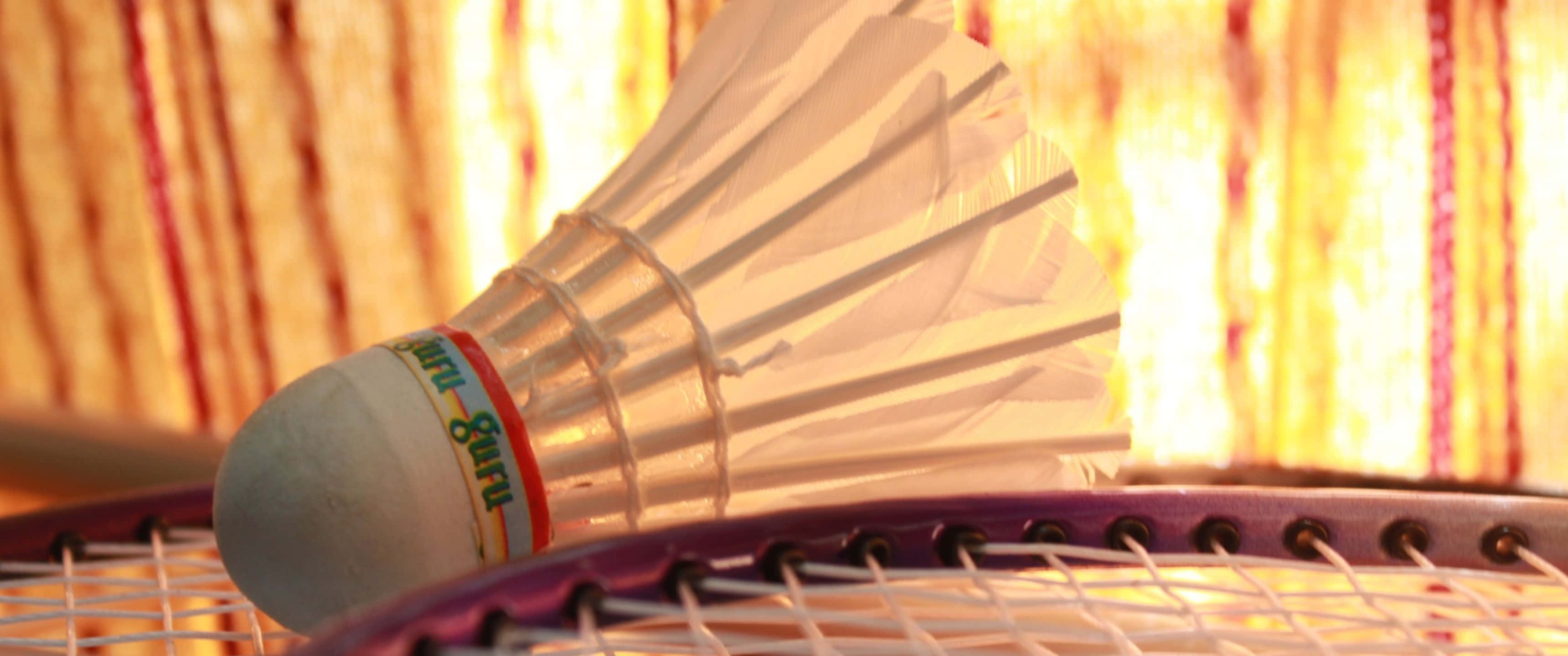 以 3440x1440p 羽球背景提升你的羽球技巧！ 壁紙 — 一個白色羽毛球嘅特寫，放在紫色羽毛球拍上，背景係溫暖嘅金色條紋。