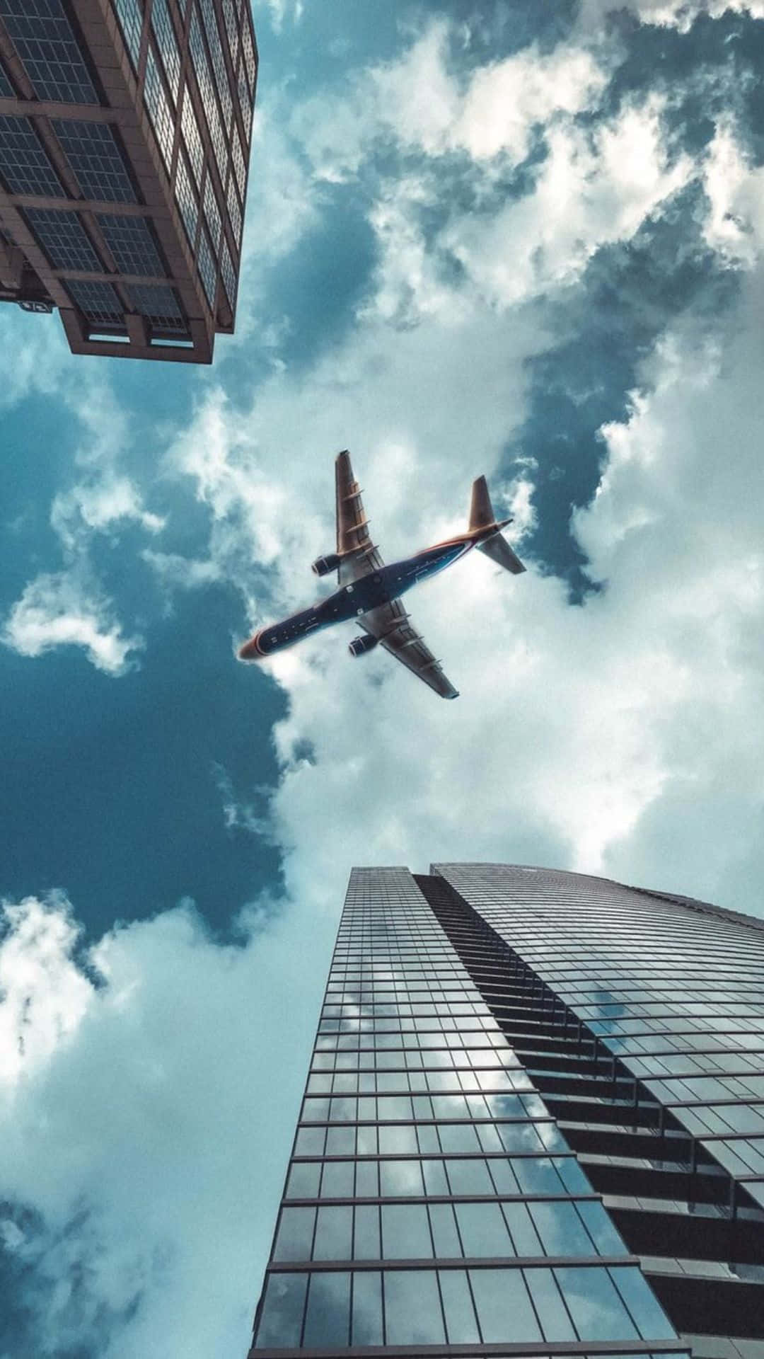 以 Android 小型飛機，時尚飛行 壁紙 — 一架商用飛機在戲劇性的多雲天空下，翱翔於高聳的玻璃摩天大樓之間。