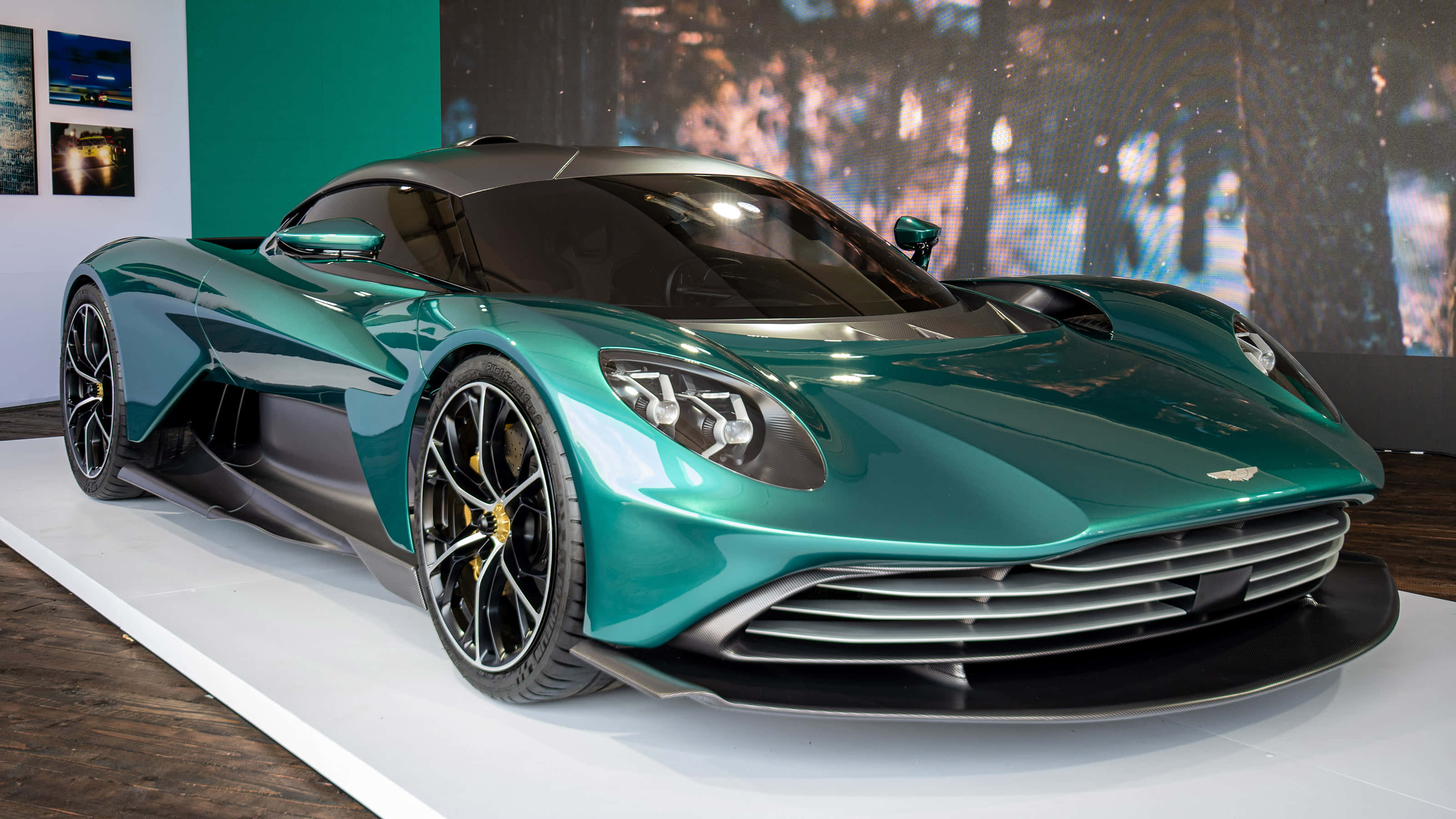 以 Aston Martin 駕馭道路風采 壁紙 — 一架時尚的藍綠色 Aston Martin 超級跑車在現代展廳的白色展示台上，在明亮燈光下閃閃發光。