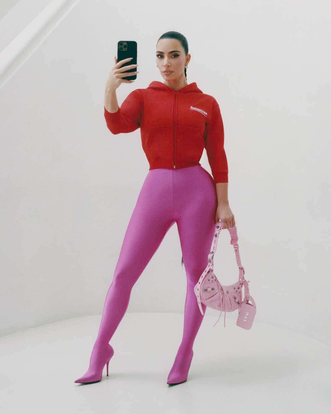 以 Balenciaga 升級衣櫥 壁紙 — 一位身穿紅色連帽衫和洋紅色緊身褲的女性，在簡約的白色工作室中拿著粉紅色手提包自拍。