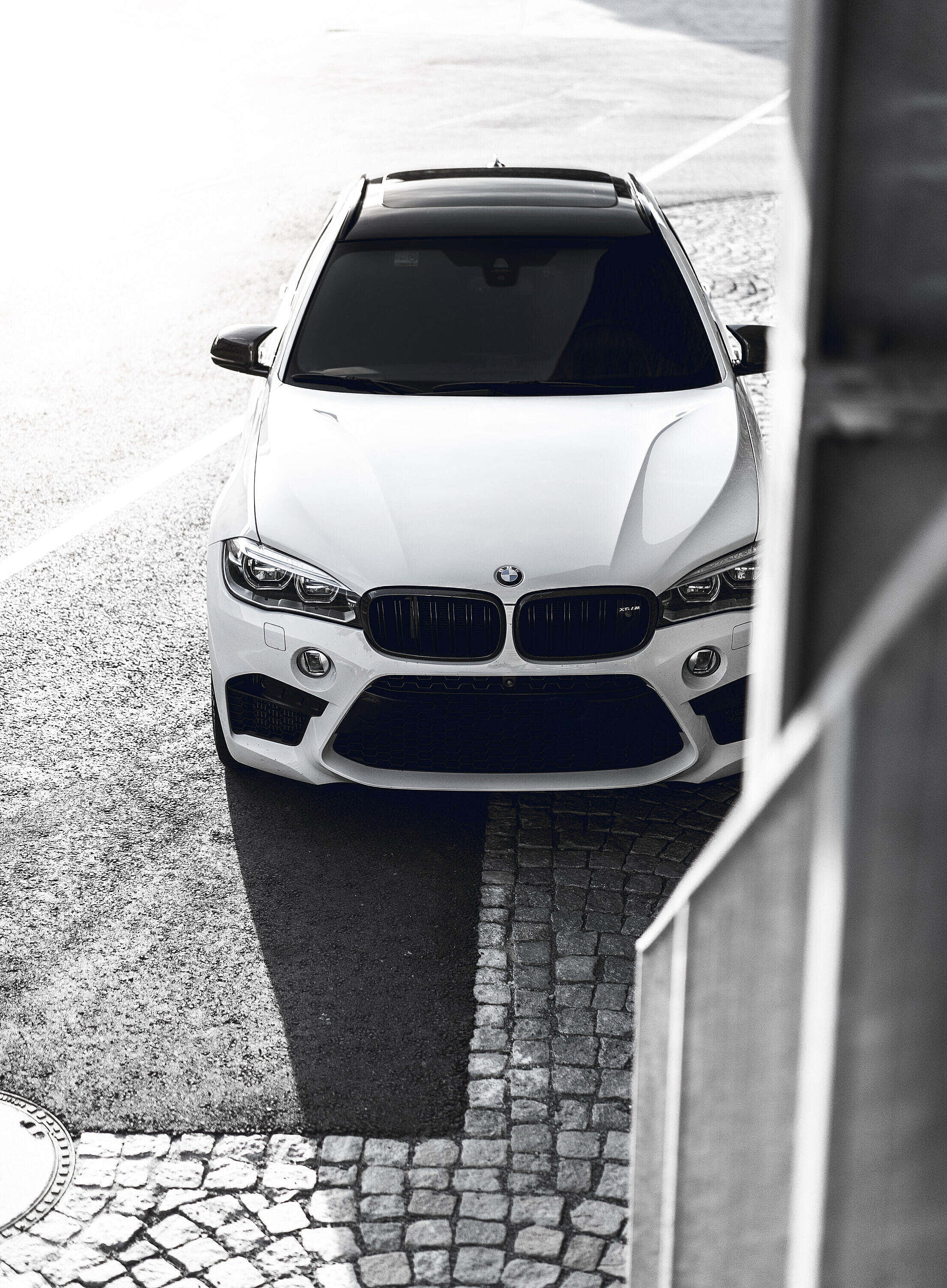 以 BMW X6 M 為榮，更強更優雅 壁紙 — 一輛時尚的白色BMW SUV從上方拍攝，位於鵝卵石街道上，帶有強烈陰影和現代建築框架。