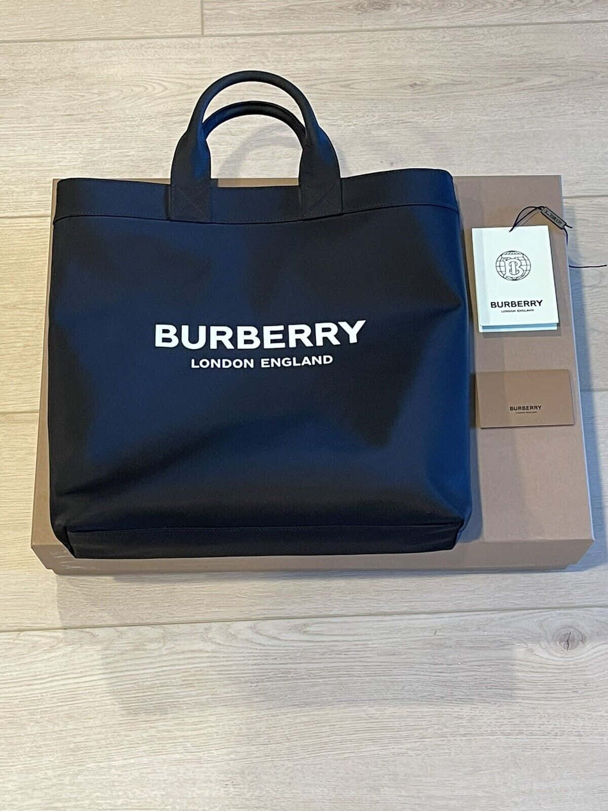 以 Burberry 展現經典英國時尚 壁紙 — 一個時尚的黑色 Burberry 手提袋，帶有白色標誌，放在其品牌盒子上，位於淺色木地板上。