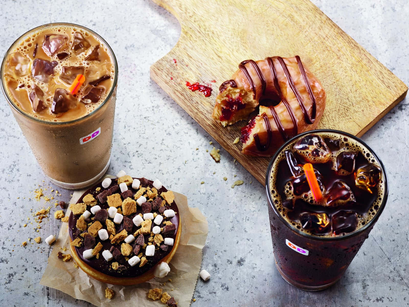 以 Dunkin' Donuts 甜甜圈犒賞自己 壁紙 — 兩杯冰咖啡飲品配以朱古力和橙色的飲管，旁邊放著一個朱古力包裹的紅桑子餡冬甩和一個烤棉花糖夾心餅風格的冬甩，置於牛油紙上。