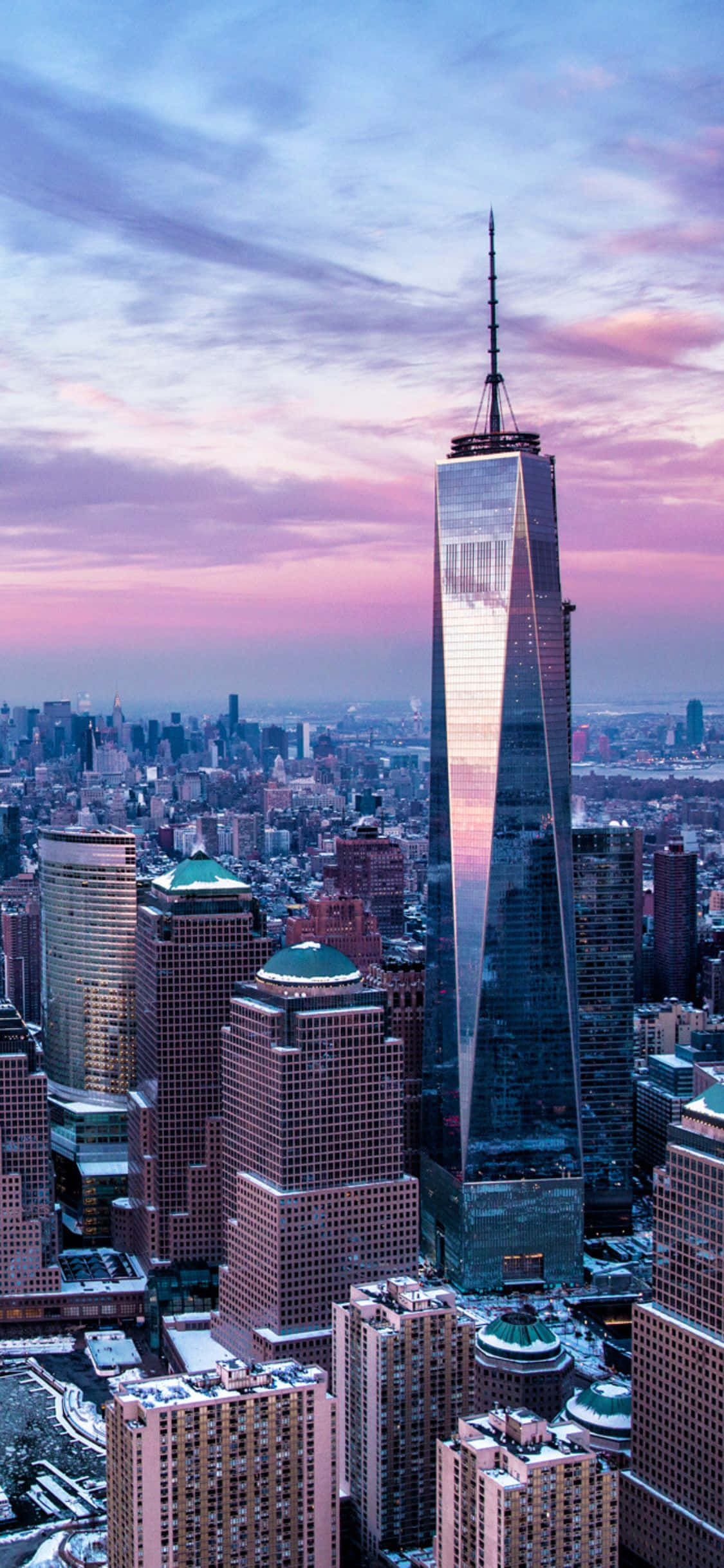 以 iPhone X 欣賞紐約天際線的美景 壁紙 — 一座高聳的玻璃幕牆摩天大樓，在充滿活力的城市天際線中反射出粉紅色和紫色的日落色調。