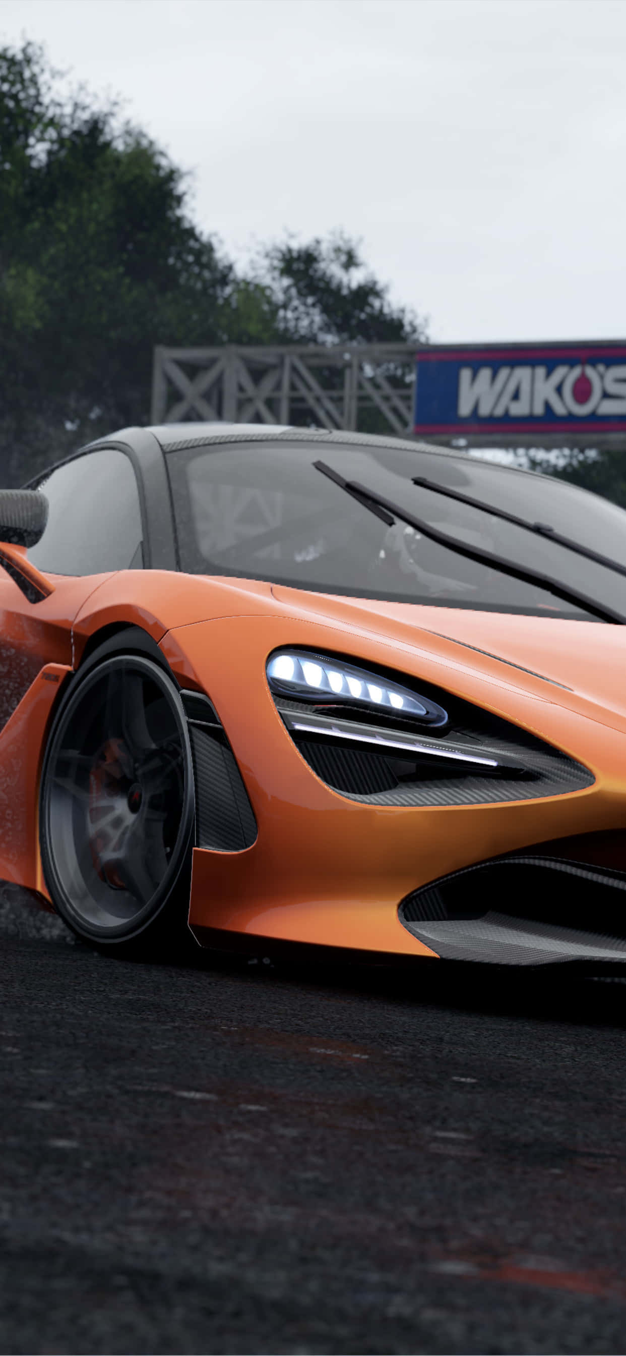 以 iPhone XS Max 沉浸於 Project Cars 2 的高速世界 壁紙 — 一輛時尚的橙色McLaren跑車，車頭燈發光，停泊在潮濕的賽道上，天空多雲。